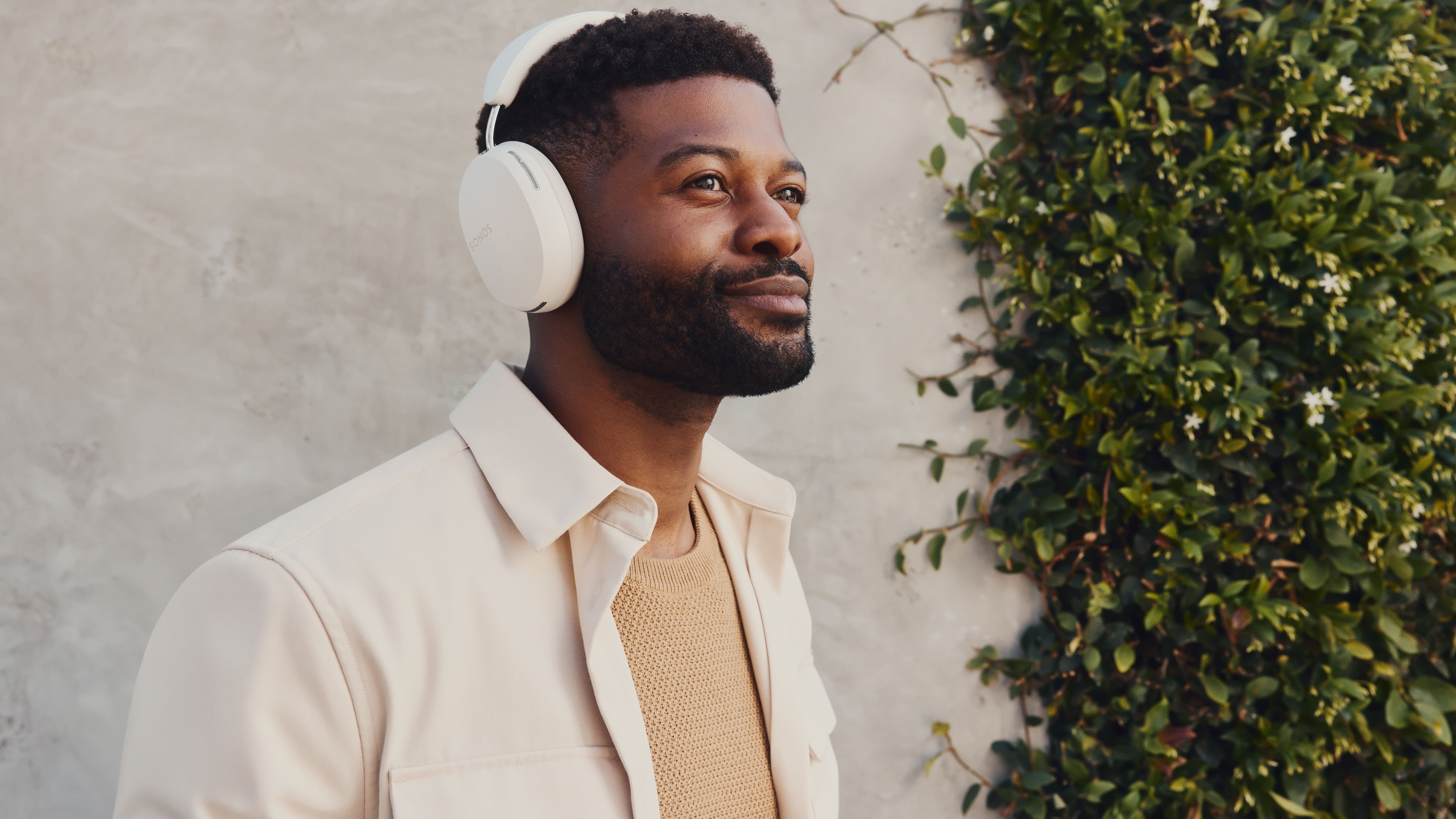 A pair of white Sonos Ace being worn by a man in front of a white wall with a vine growing up it.