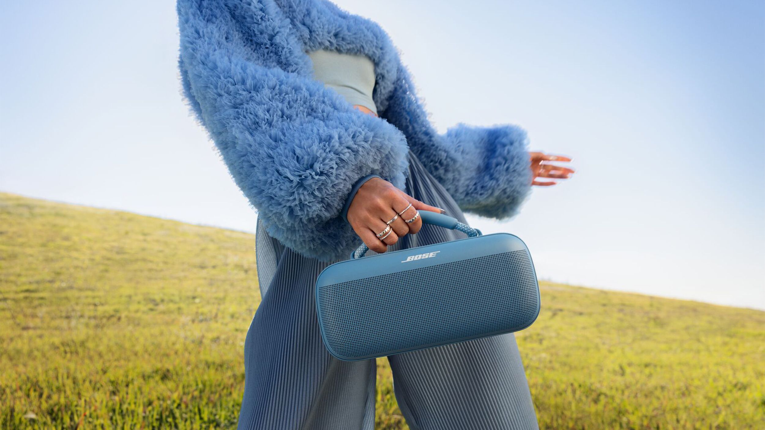 A person wearing a blue fuzzy top holds the blue Bose SoundLink Max speaker in front of a grassy hill and blue sky.