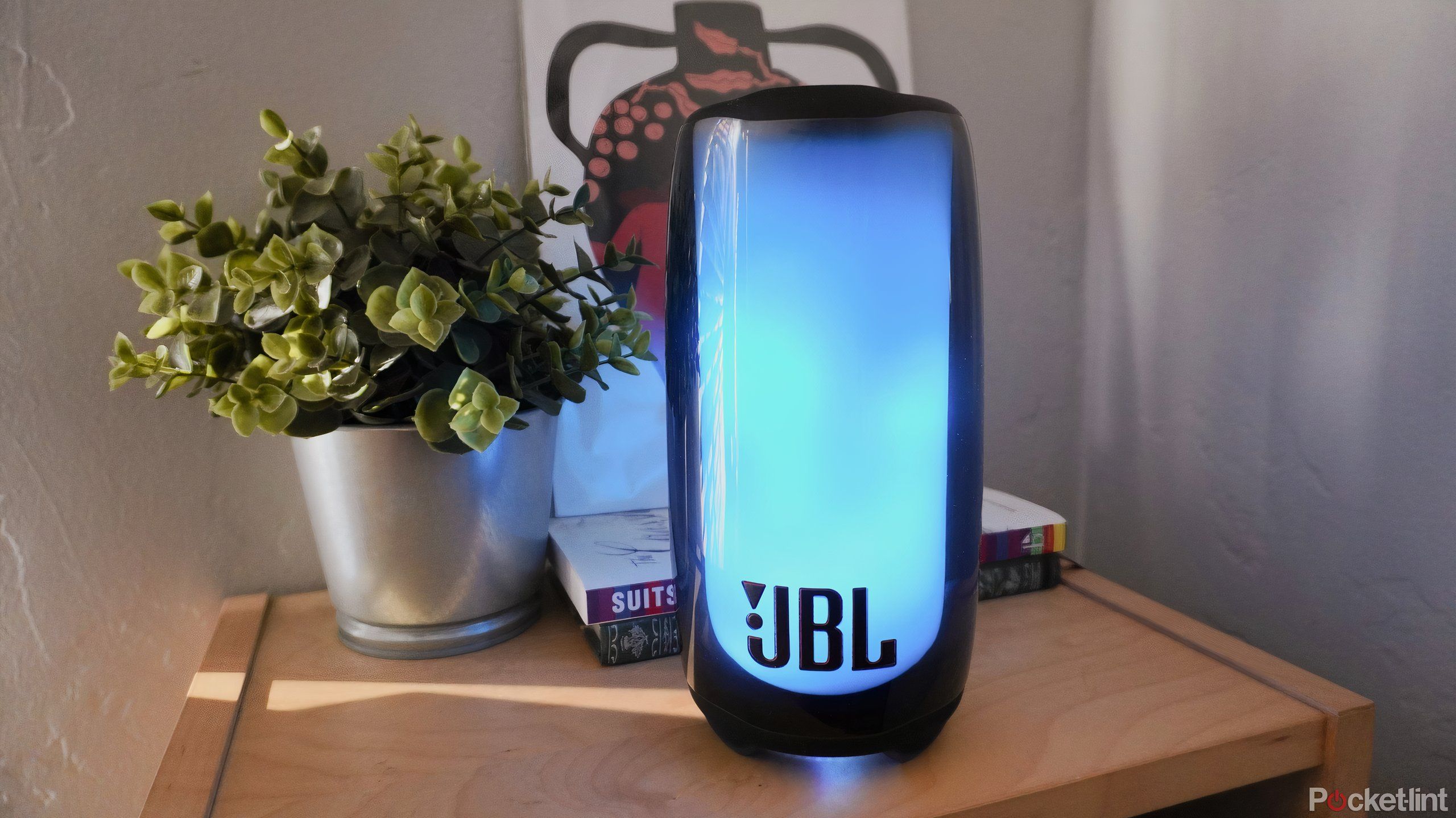 A glowing JBL Pulse 5 speaker on a wooden bookshelf.
