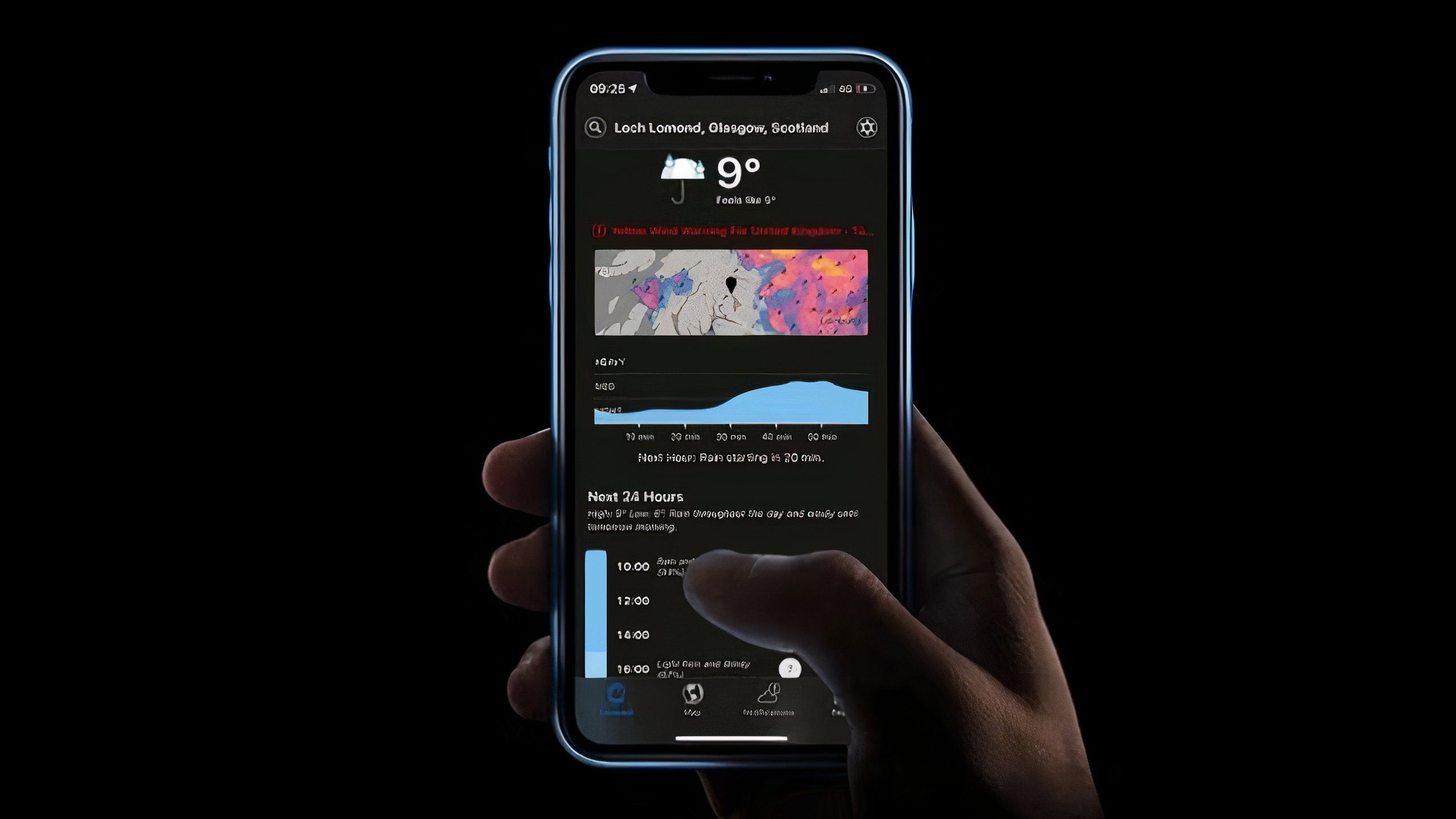 A hand holding an iPhone 11 running the now discontinued Dark Sky weather app.