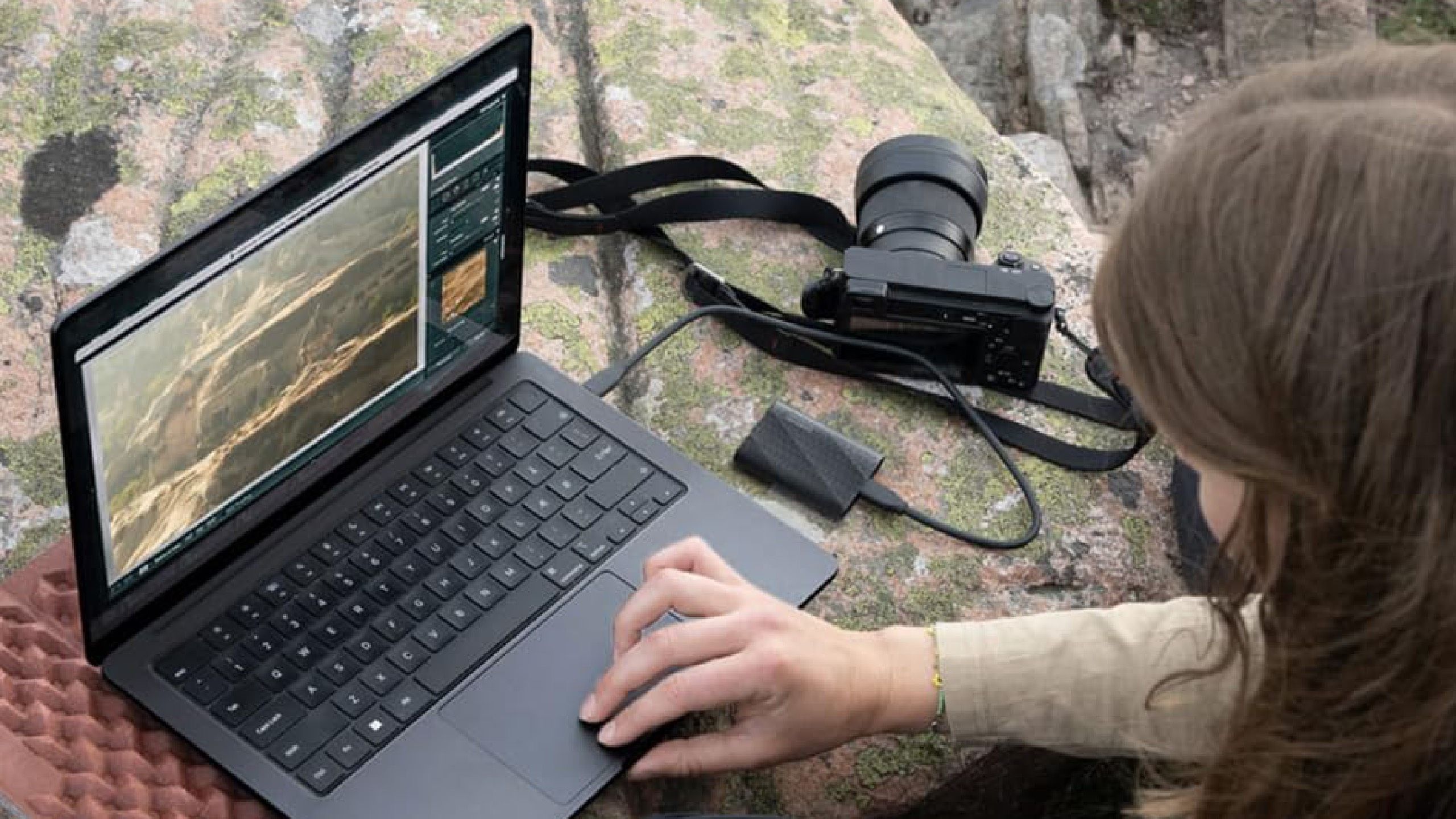 A person using their computer outside on a mossy rock. 