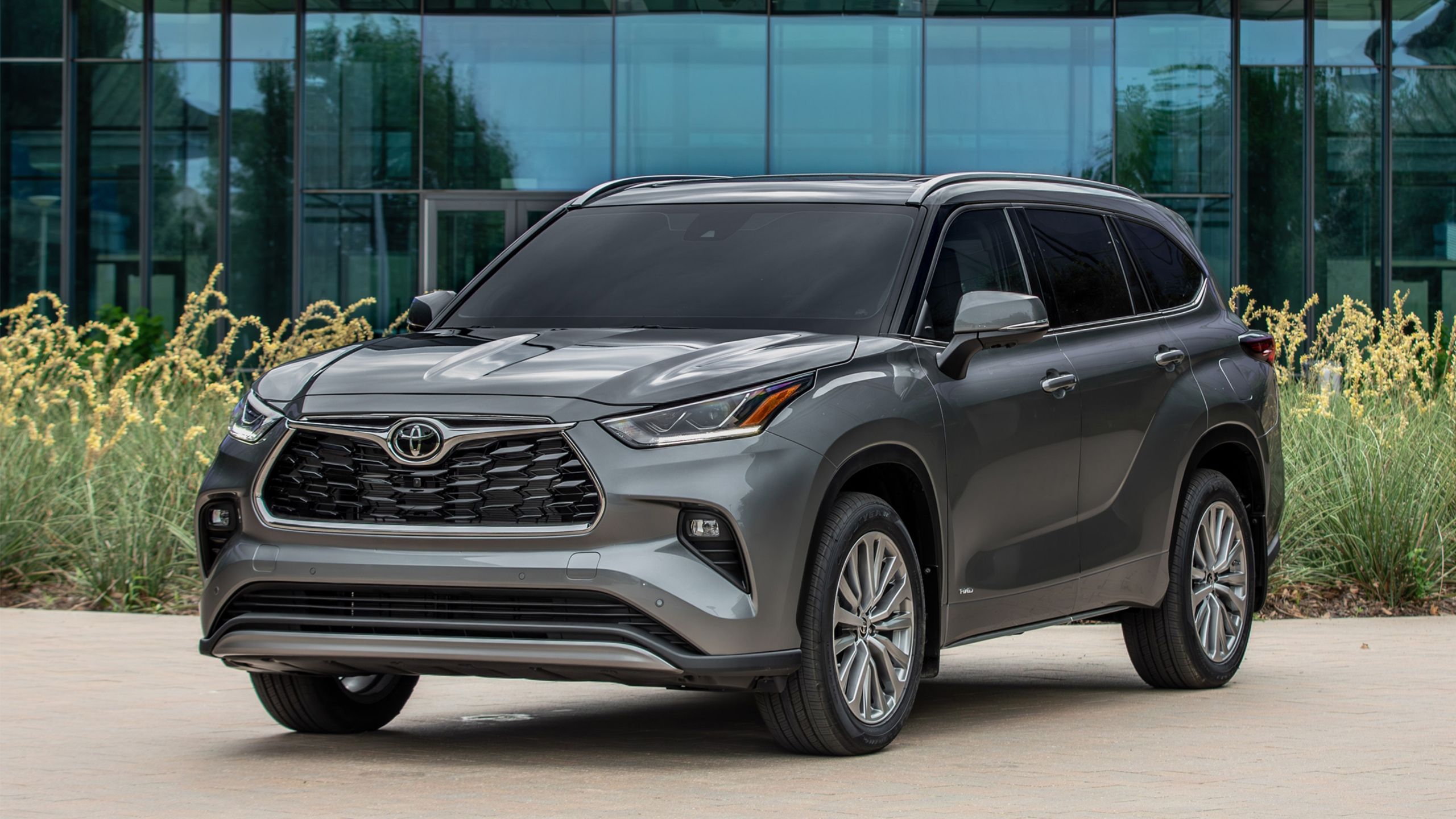 Front quarter view of the 2025 Toyota Highlander hybrid parked in front of a glass building.