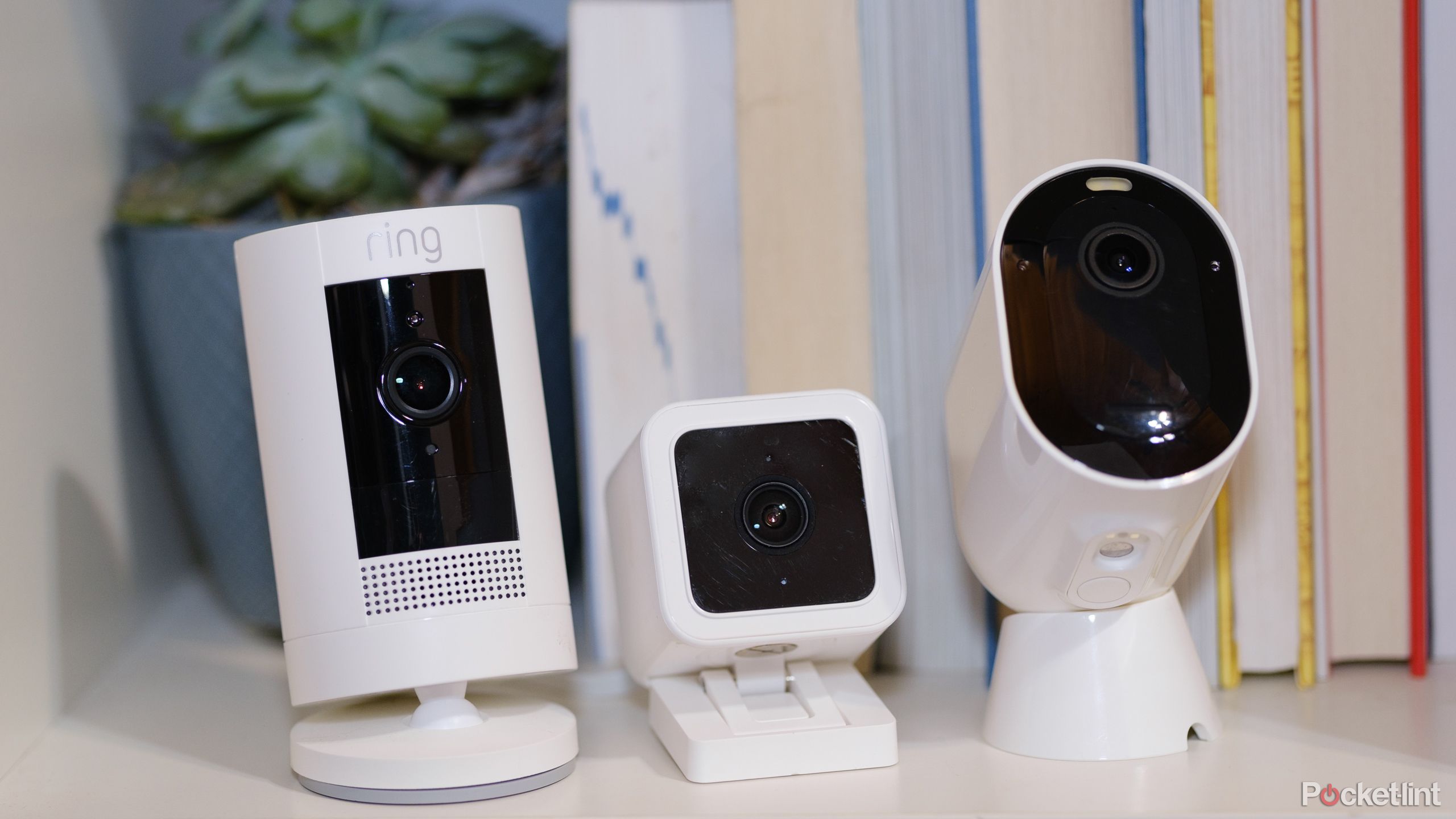 A photo of a Ring, Wyze, and Arlo security camera on a bookshelf