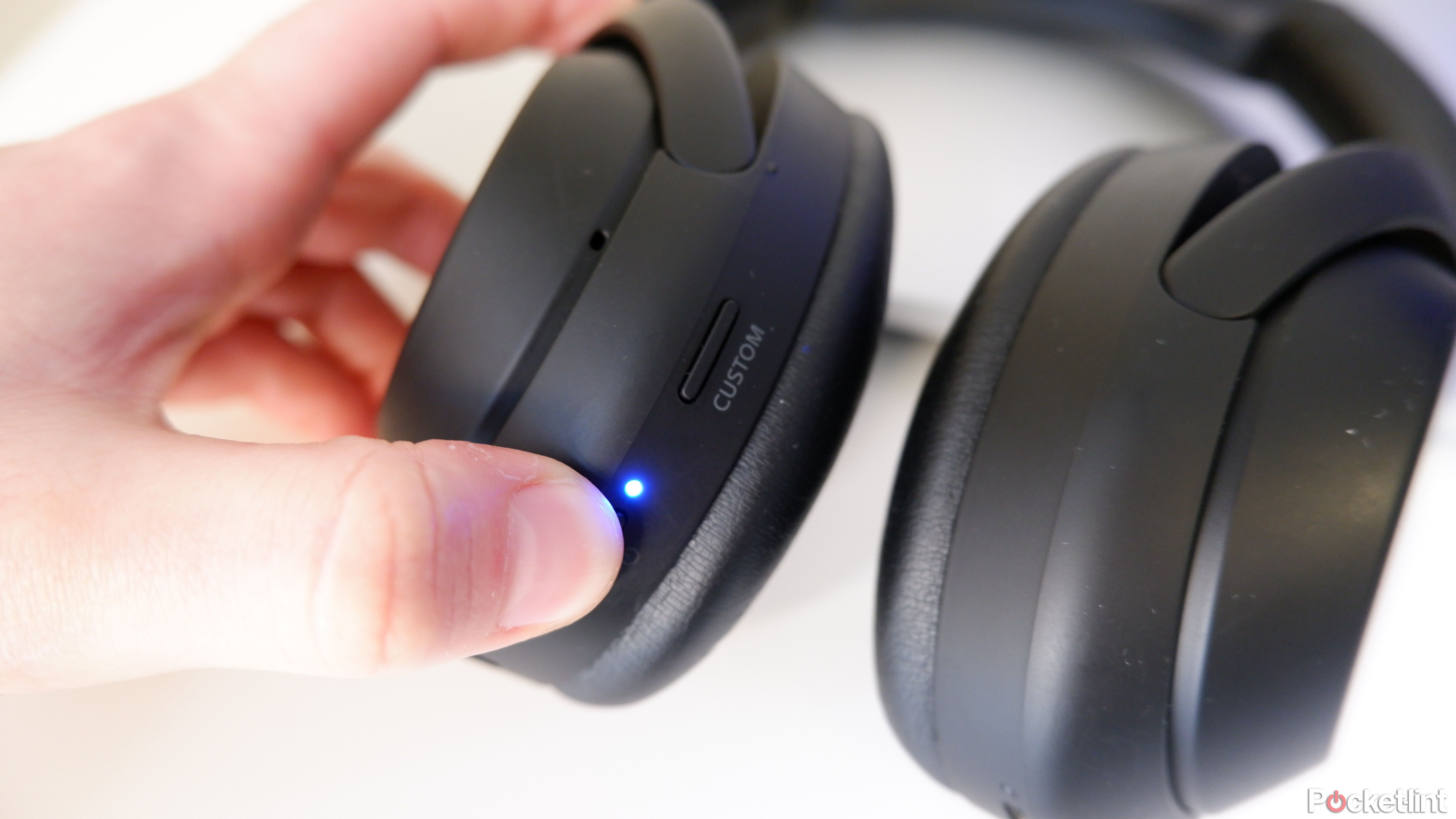 The Sony WH-1000XM4 is in Bluetooth pairing mode with your thumb pressing the power button and the blue LED illuminated.