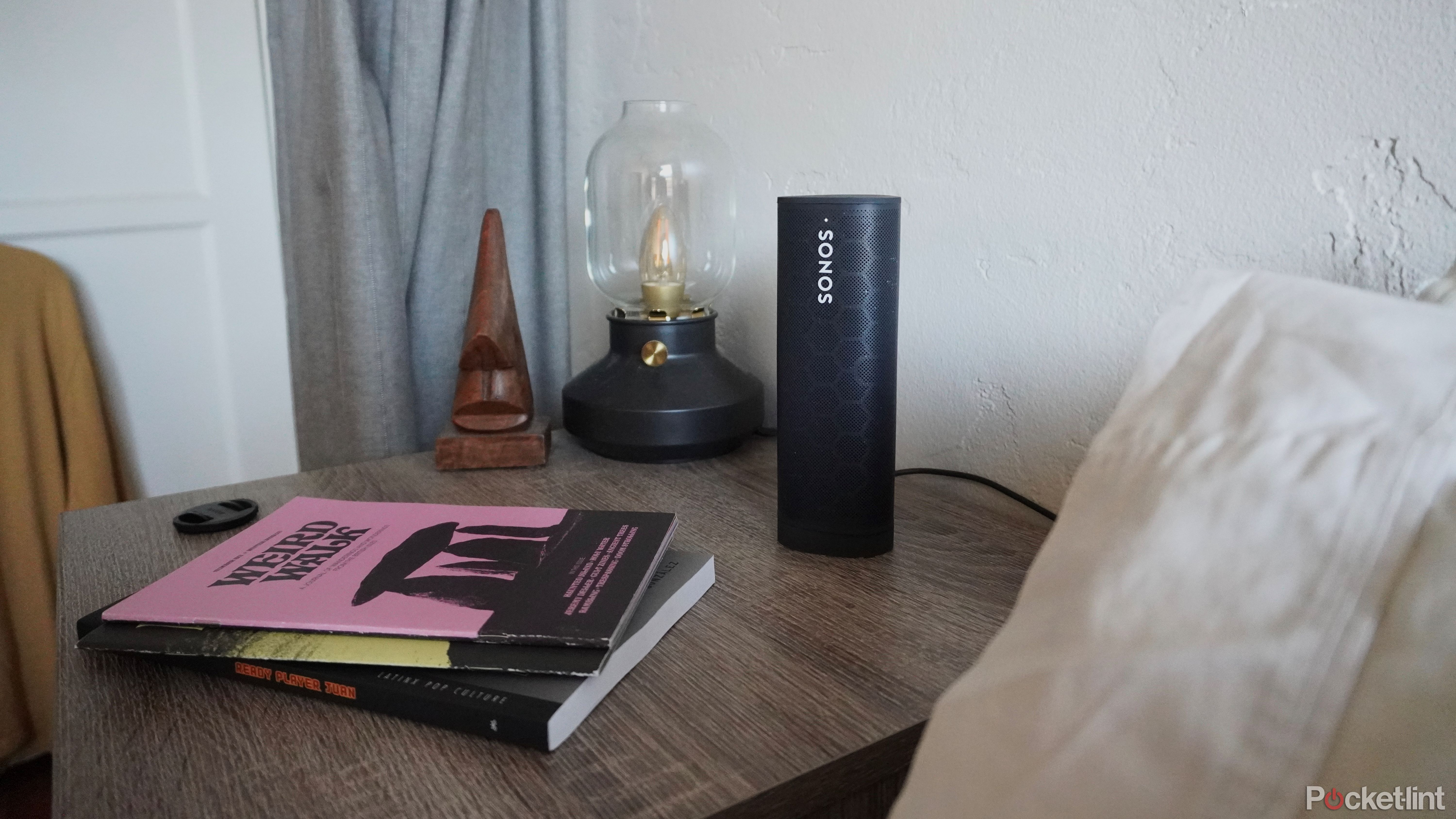 A Sonos Roam speaker on a nightstand, next to a lamp.