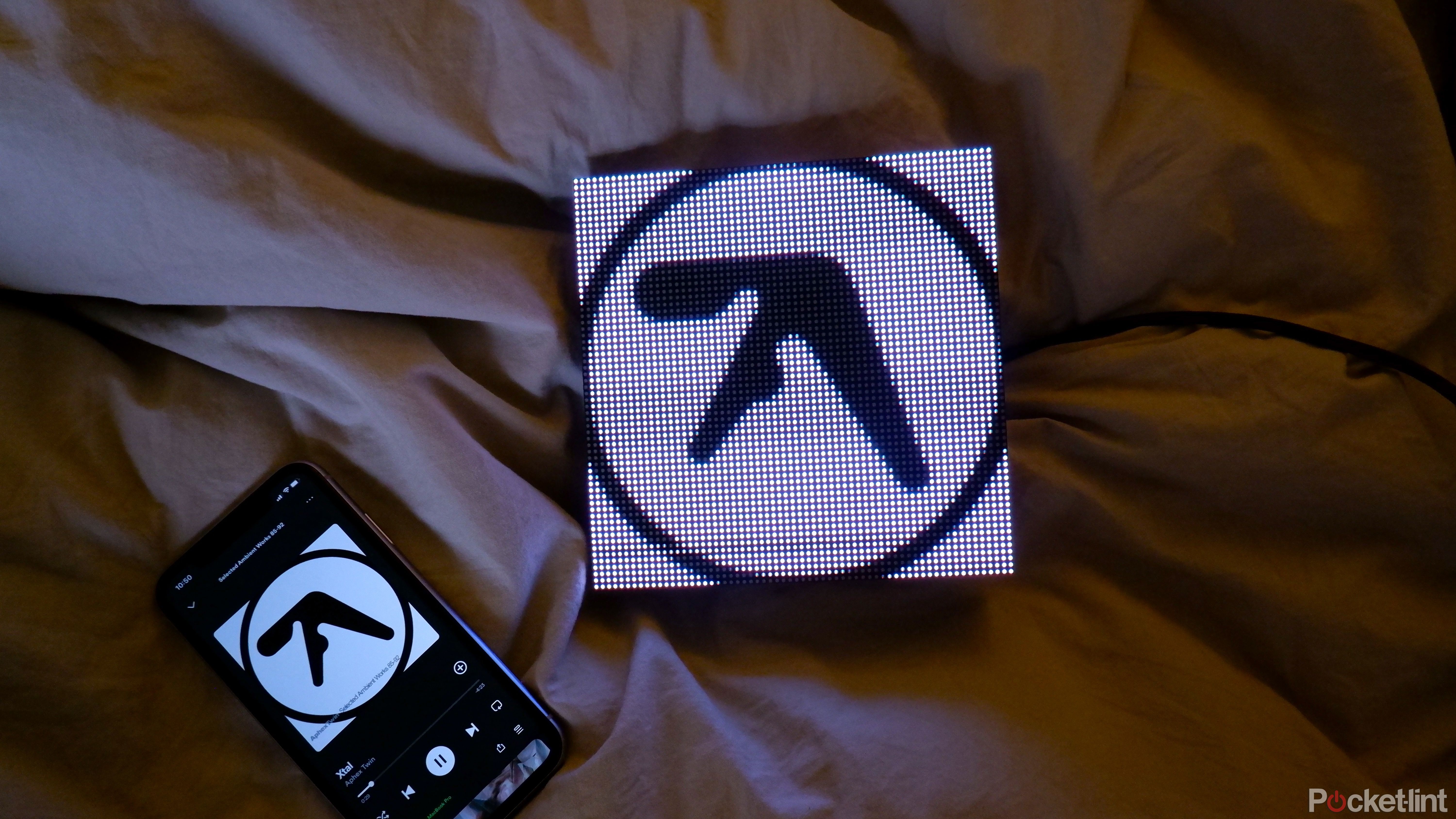 A Tuneshine on a bed showing album art for Aphex Twin, next to an iPhone with Spotify open.