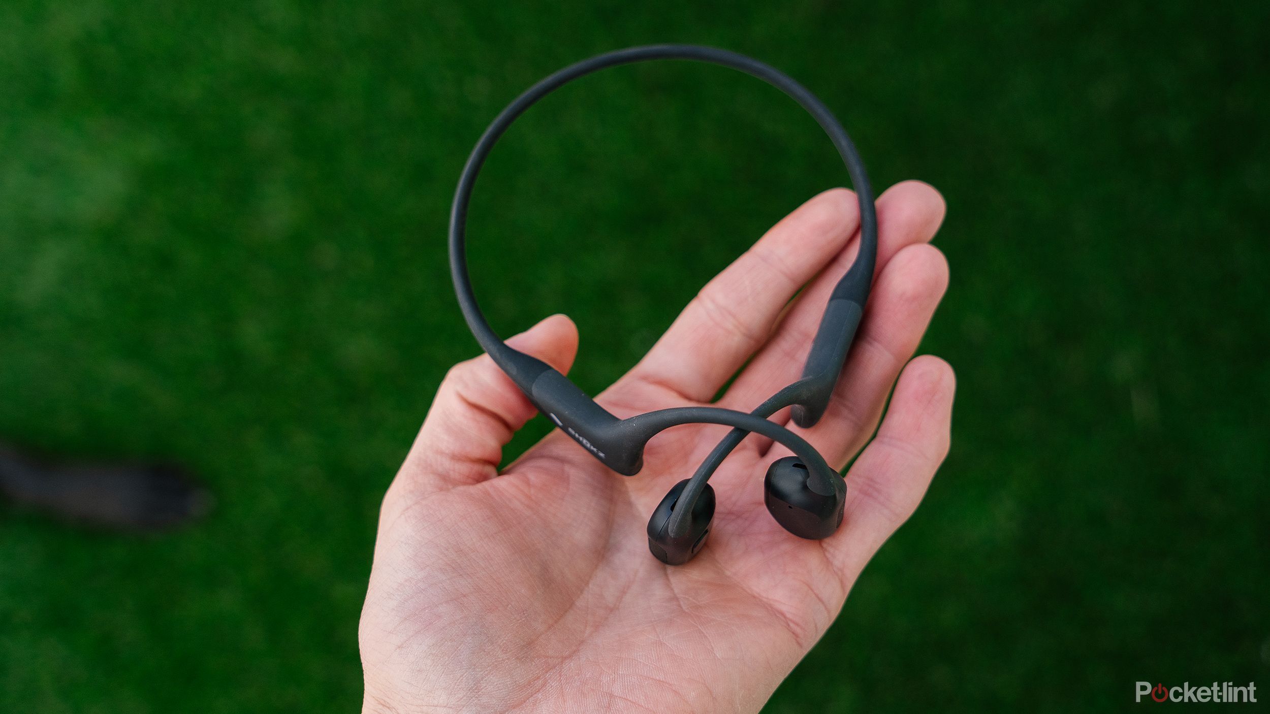 A pair of Shokz bone conduction headphones sit in a hand above blurred out grass. 