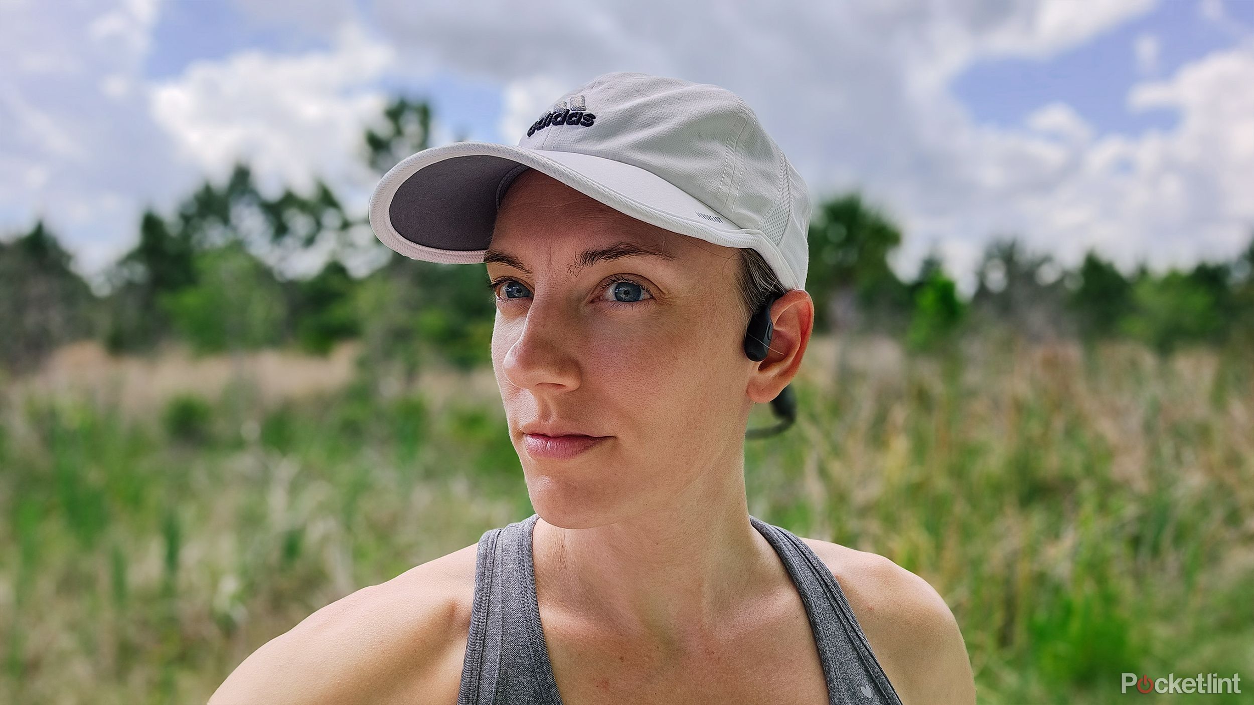 A woman with a white running hat in front of a blurred out forest wearing Shokz Open Run Pro bone conduction headphones.