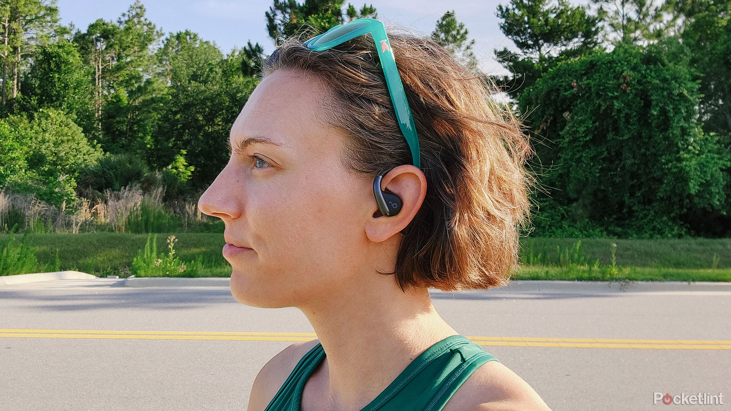 A woman stands in front of a road with forest behind it wearing the Soundcore AeroFit Open-Ear headphones.