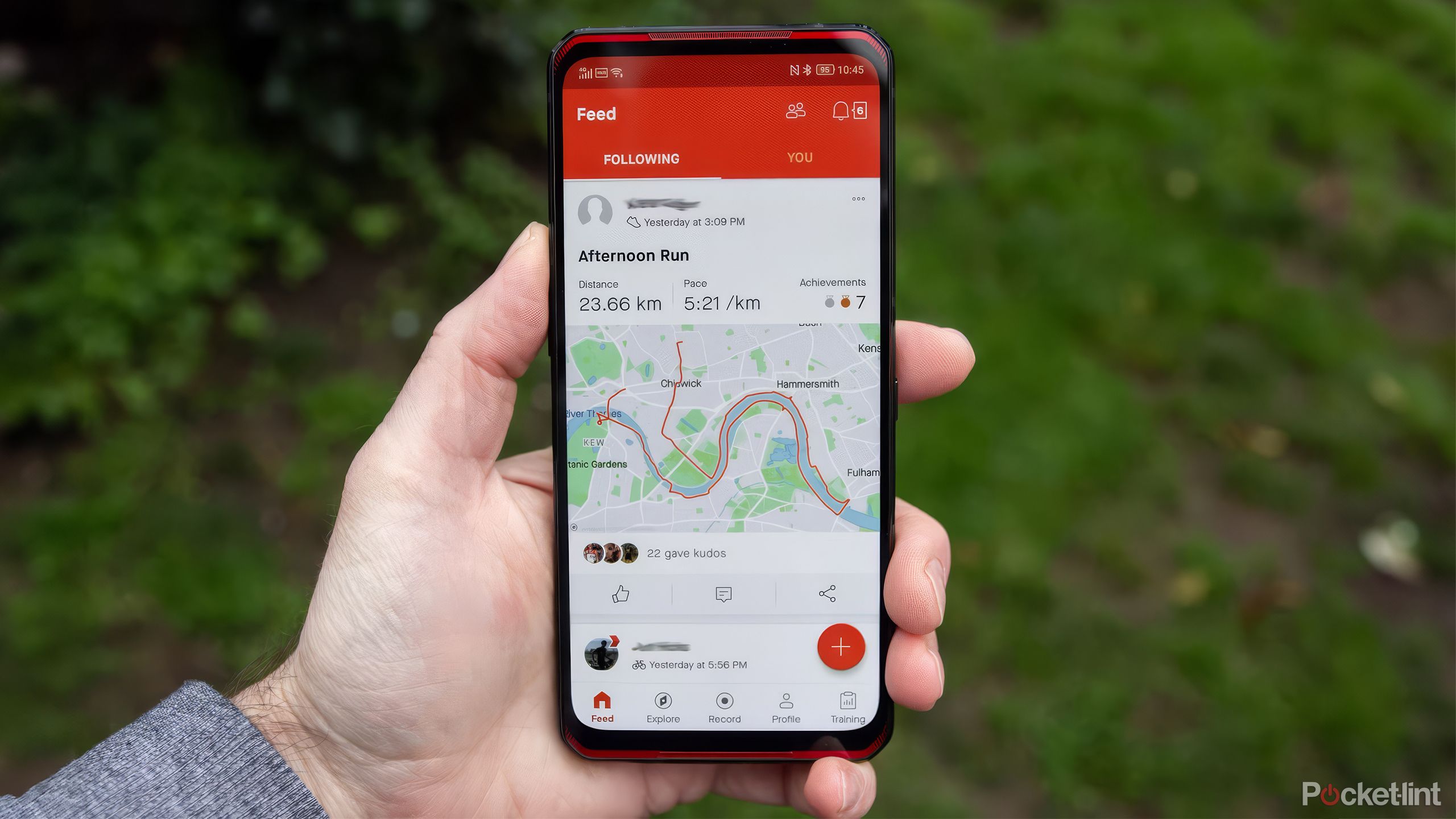 A hand holds a phone with the Strava app displayed against a blurred grassy background. 