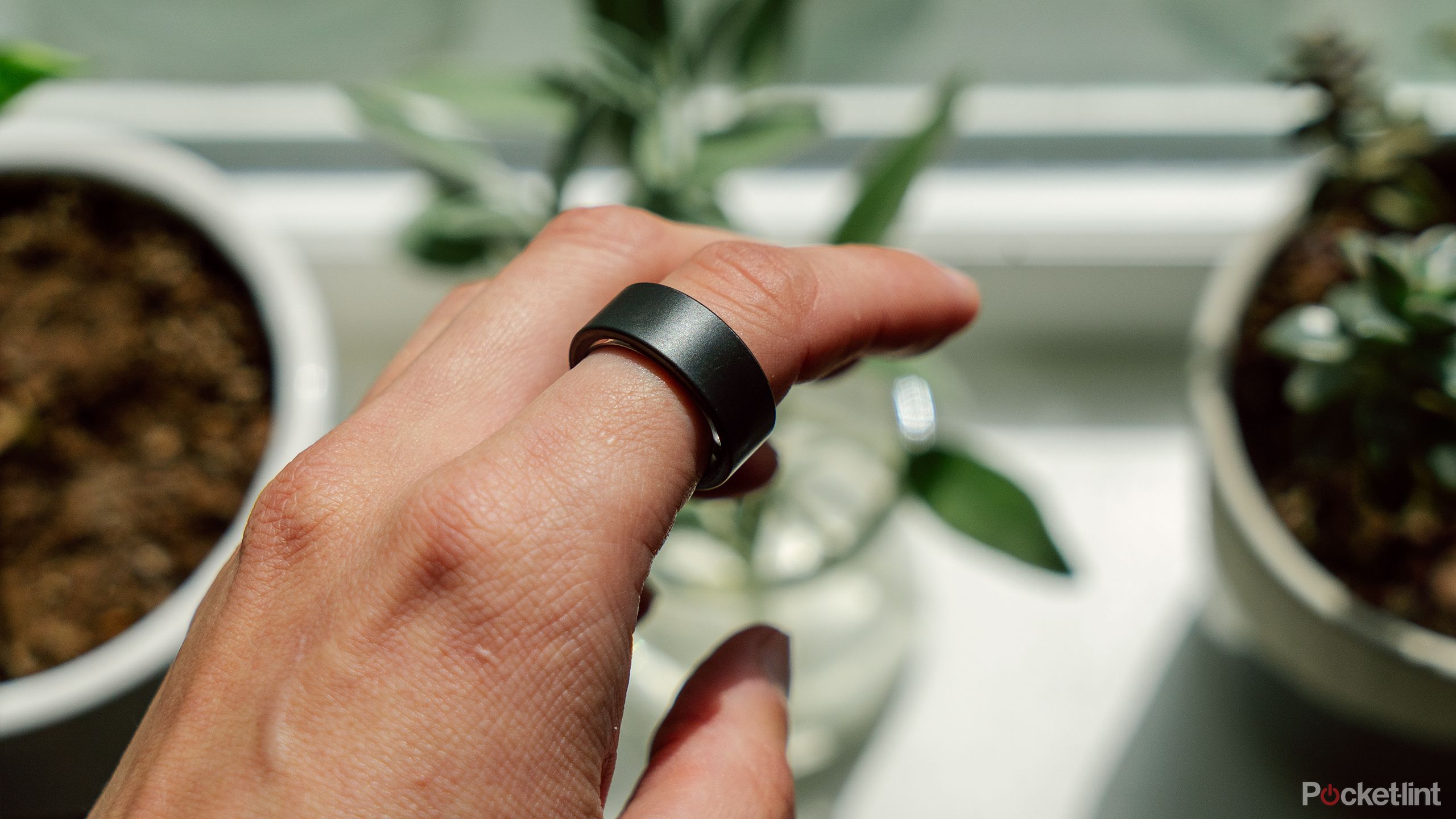 The Ultrahuman Ring Air on a finger held in front of a blurred out windowsill with plants. 