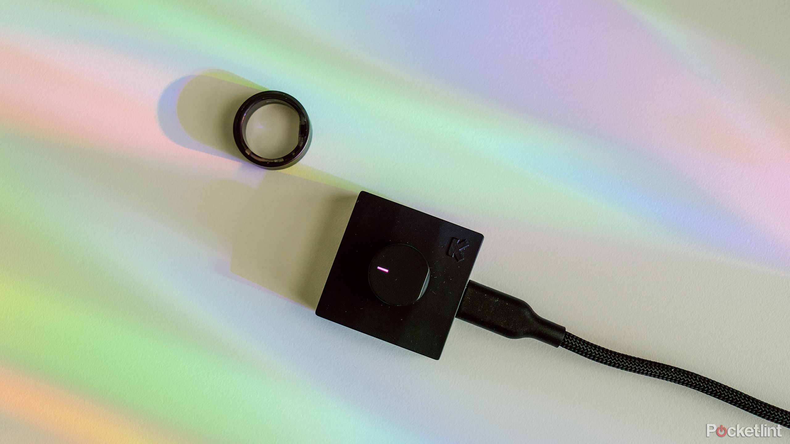 The Ultrahuman Ring Air next to its charger on a white desk with rainbow light. 
