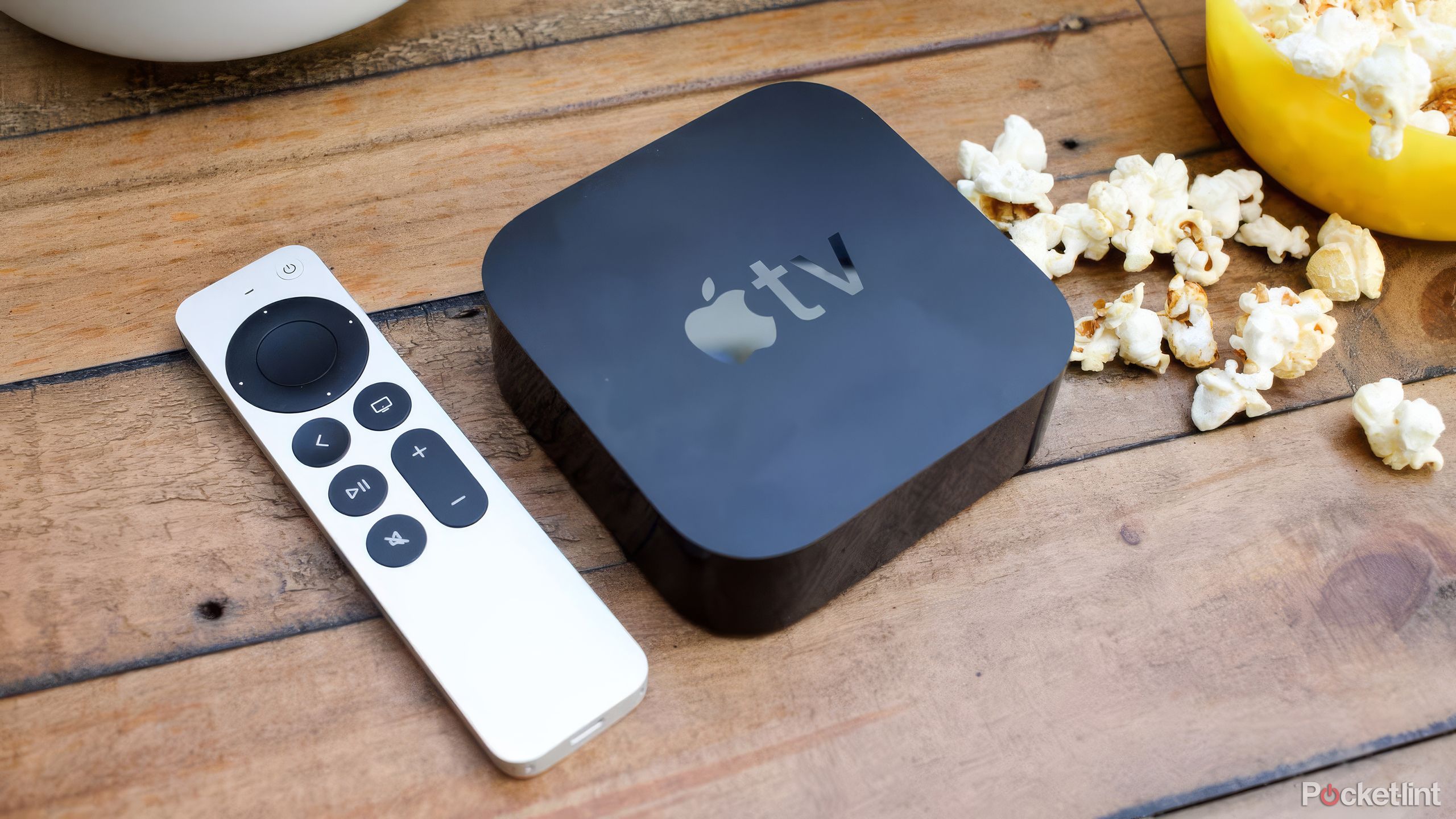 An Apple TV remote and box sit on a wood table next to popcorn.