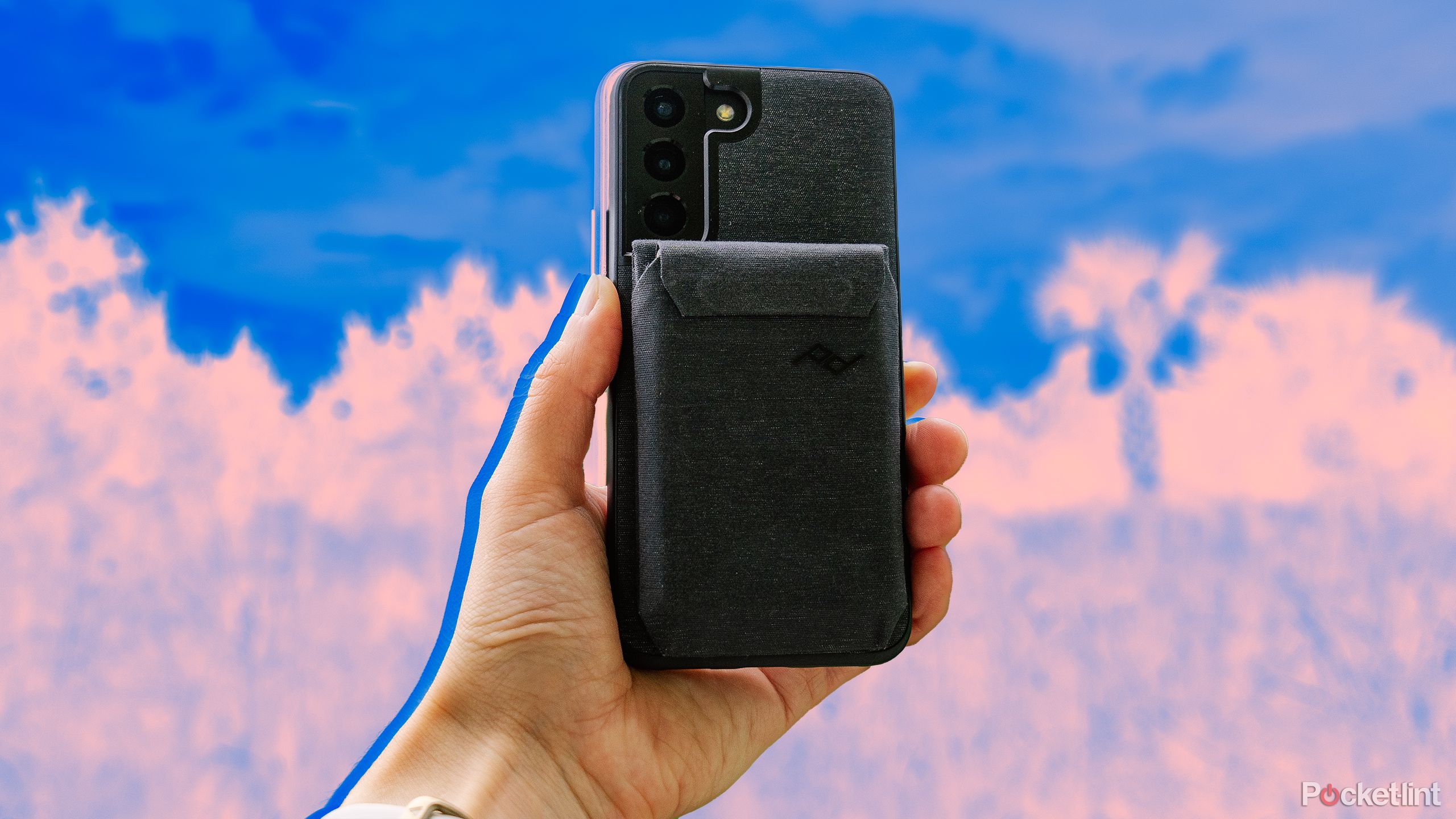 A hand holds a phone with the Peak Design Everyday Case against a blue and orange blurred background. 