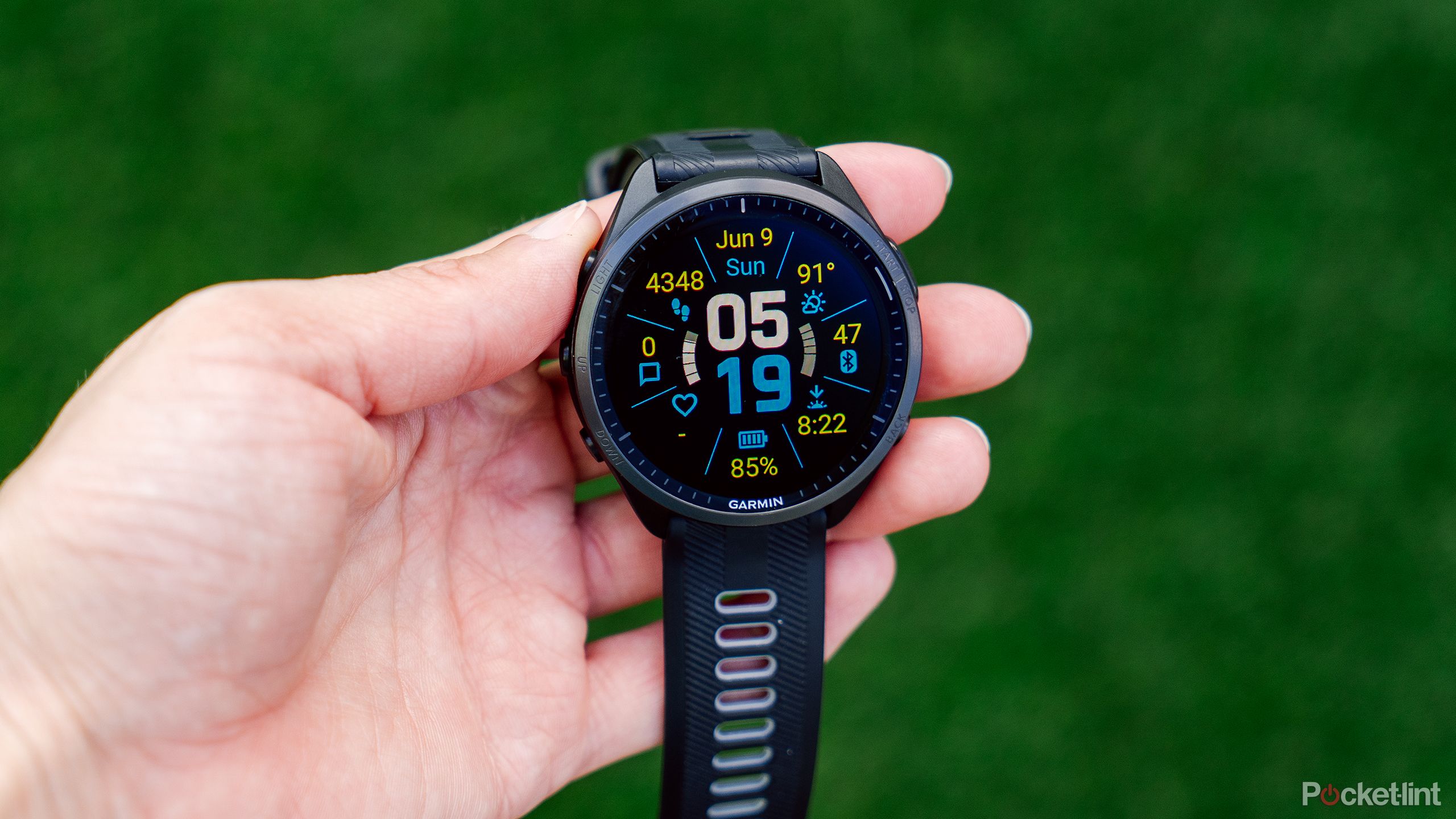 A hand holds the Garmin Forerunner 965 above a blurred green background.