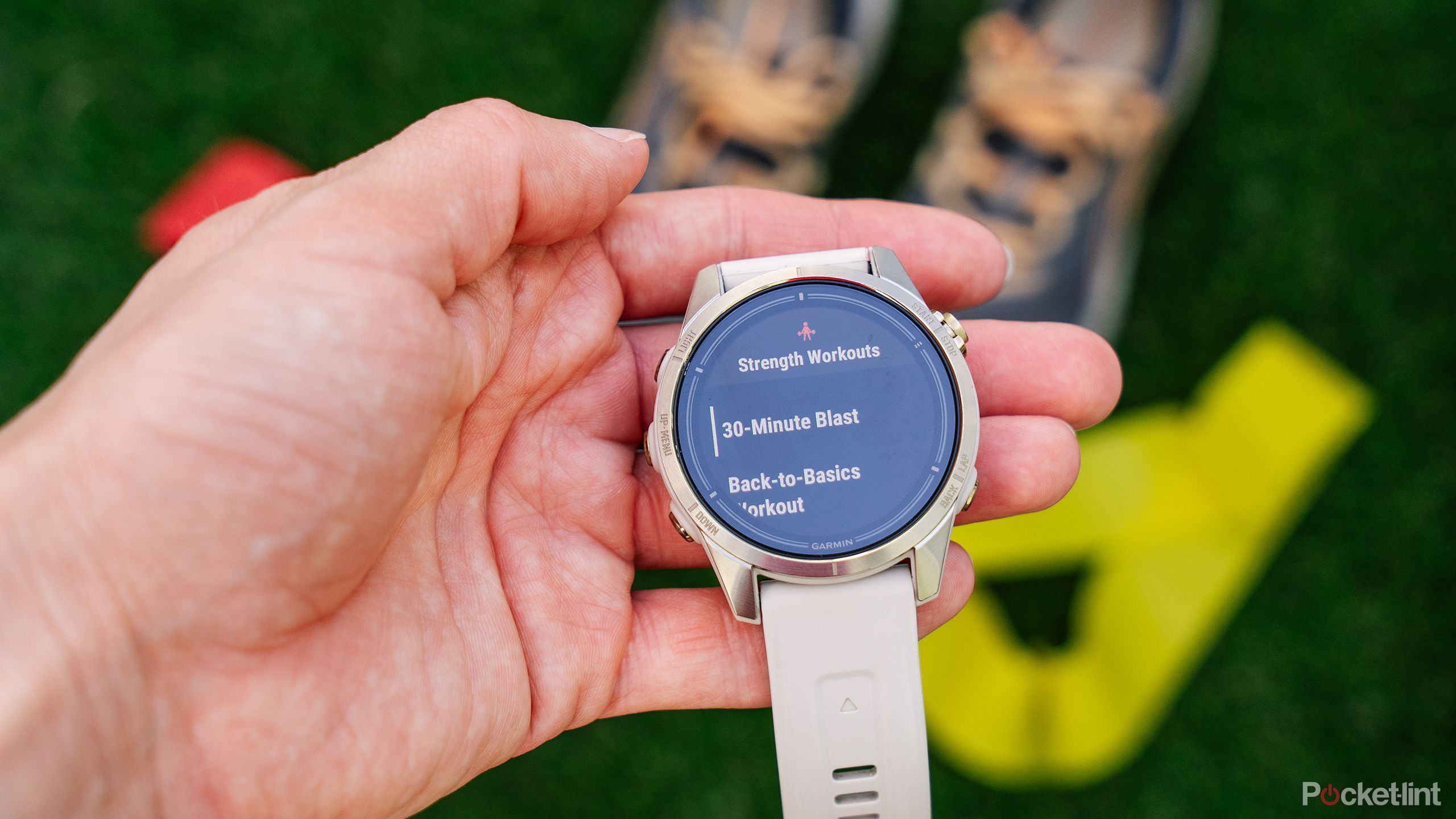 A hand holds the Garmin epix Pro Gen 2 Sapphire Edition above shoes and a yellow resistance band.