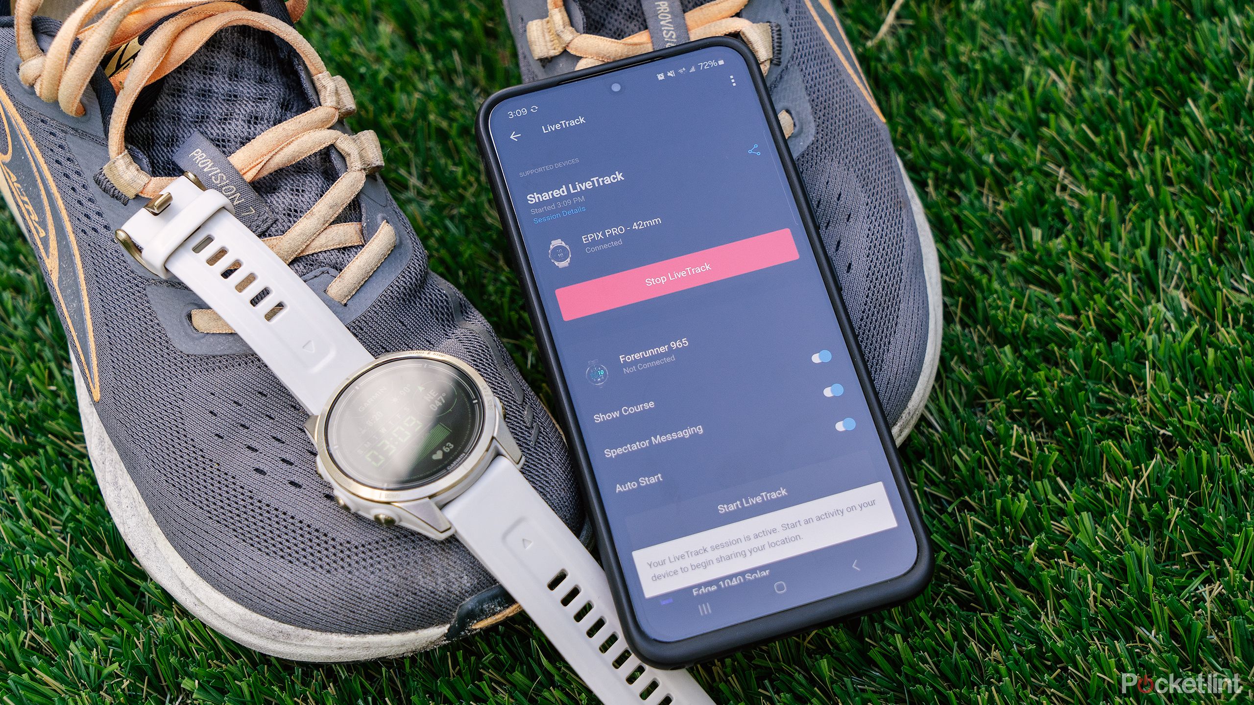 A phone with the Garmin LiveTrack feature on the screen and a Garmin watch rest on a pair of shoes on green turf.