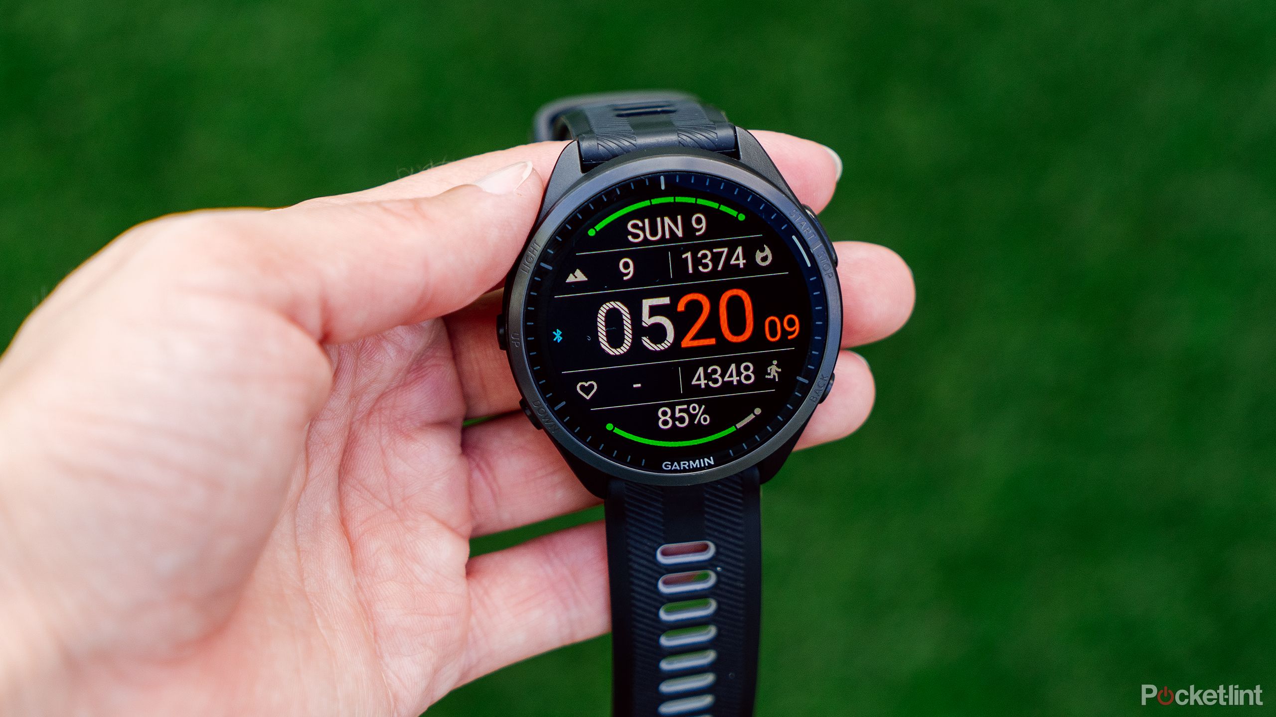 A hand holds the Garmin Forerunner 965 above a blurred green background.