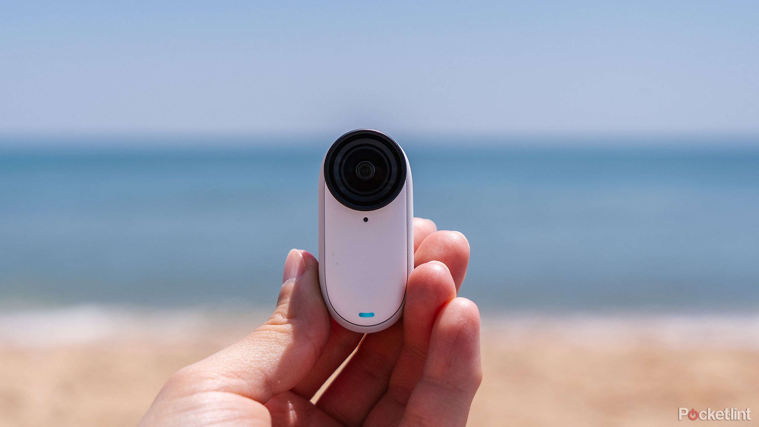 A hand holds the Insta360 GO 3S camera in front of a beach.