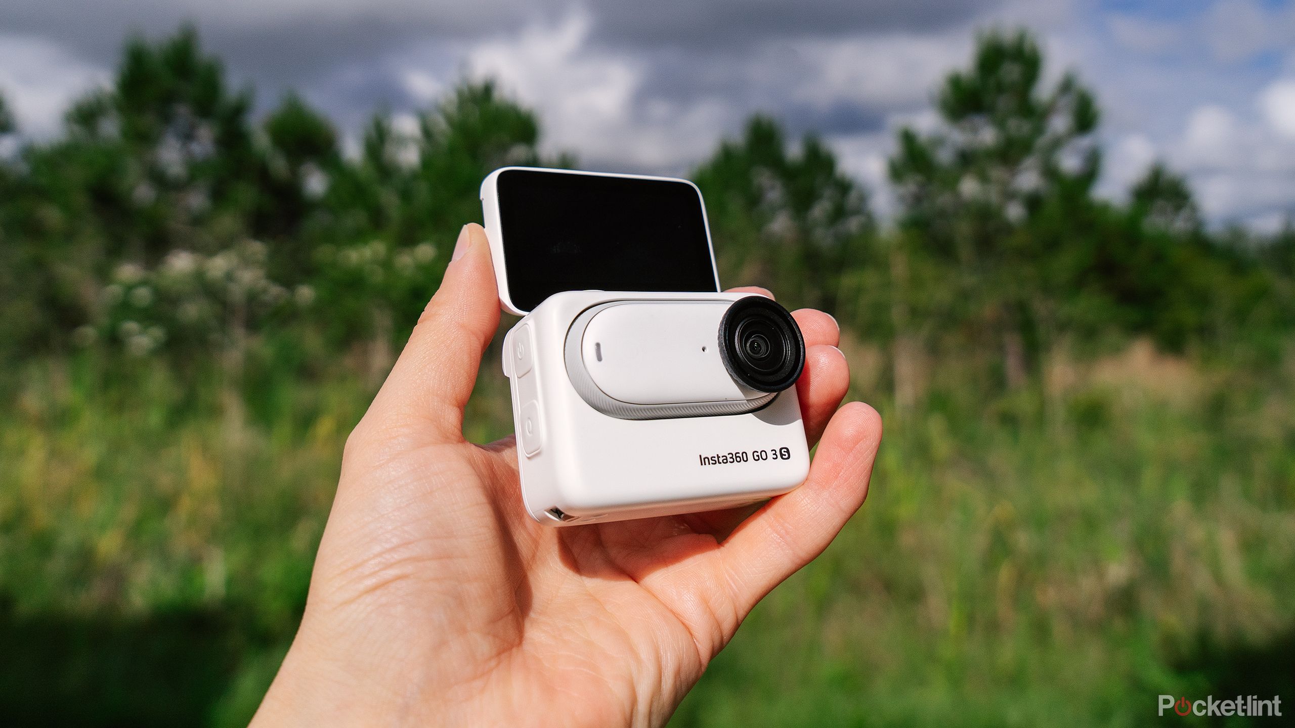 A hand holds the Insta360 GO 3S in the Action Pod with the screen flipped up. 