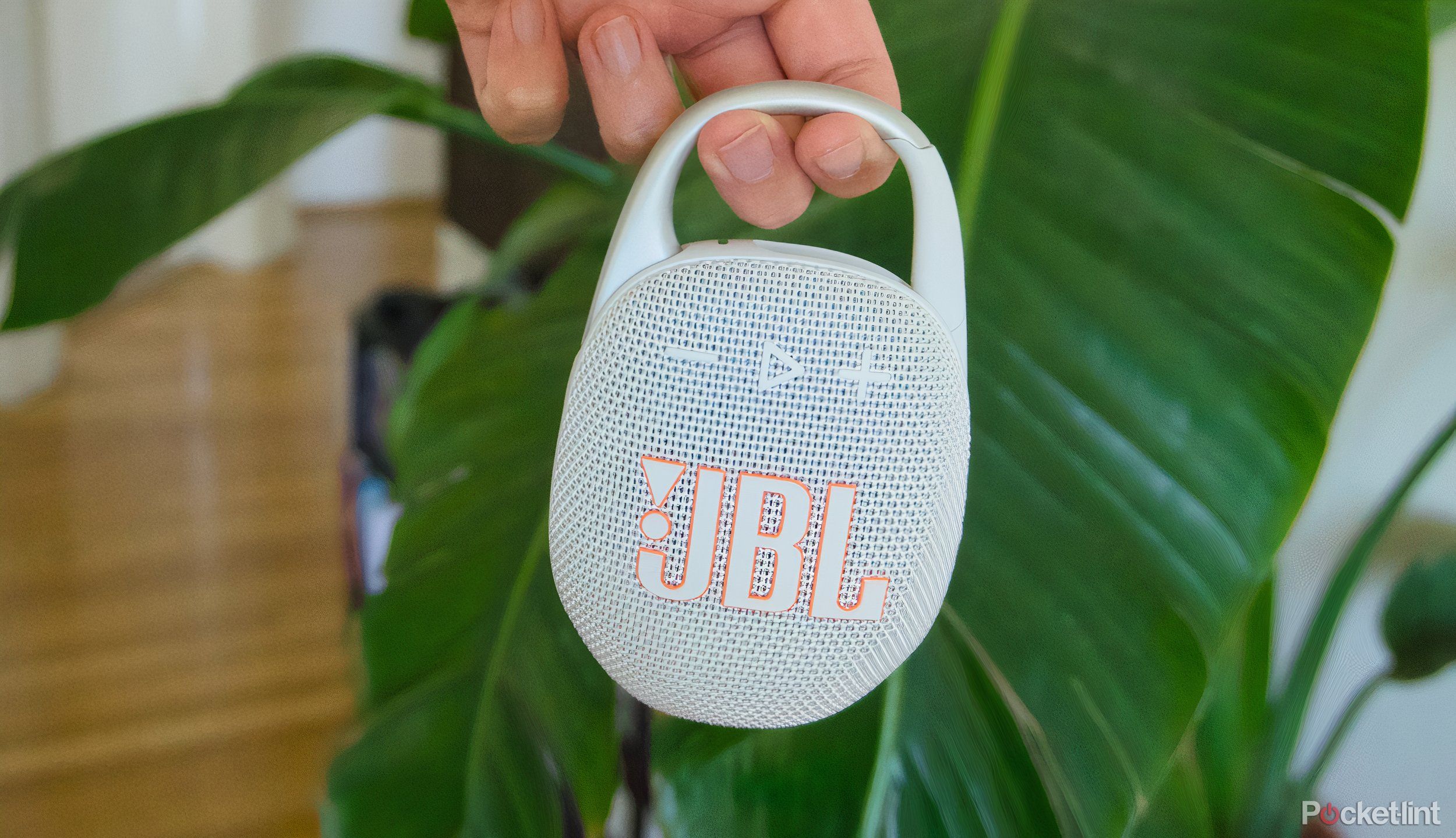 A JBL Clip 5 dangling from two fingers in front of a plant
