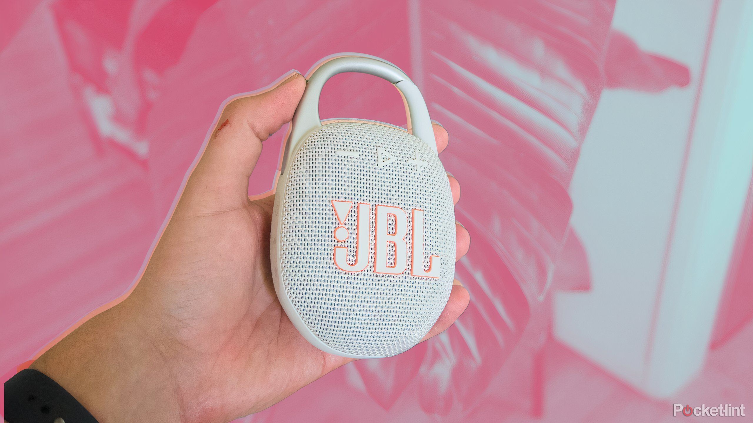 A JBL Clip 5 speaker being held in someone's hand over a pink background.
