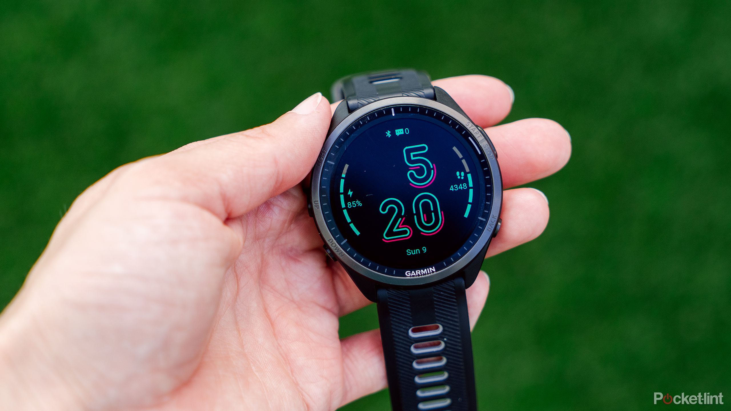 A hand holds a Garmin Forerunner 965 over a blurred green background. 