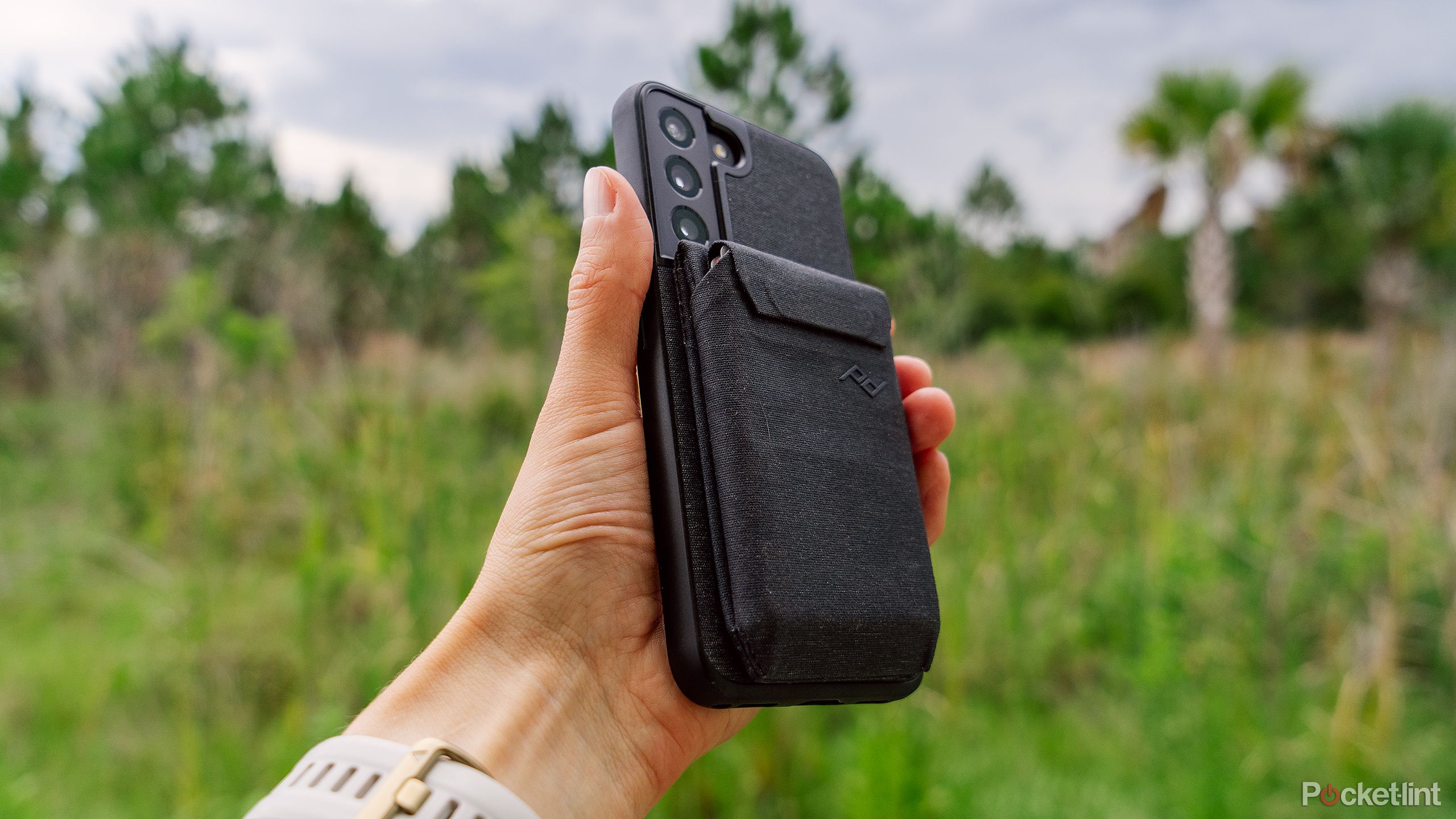 A hand holds a phone with the Peak Design Everyday Case and wallet in front of a blurred forest background. 