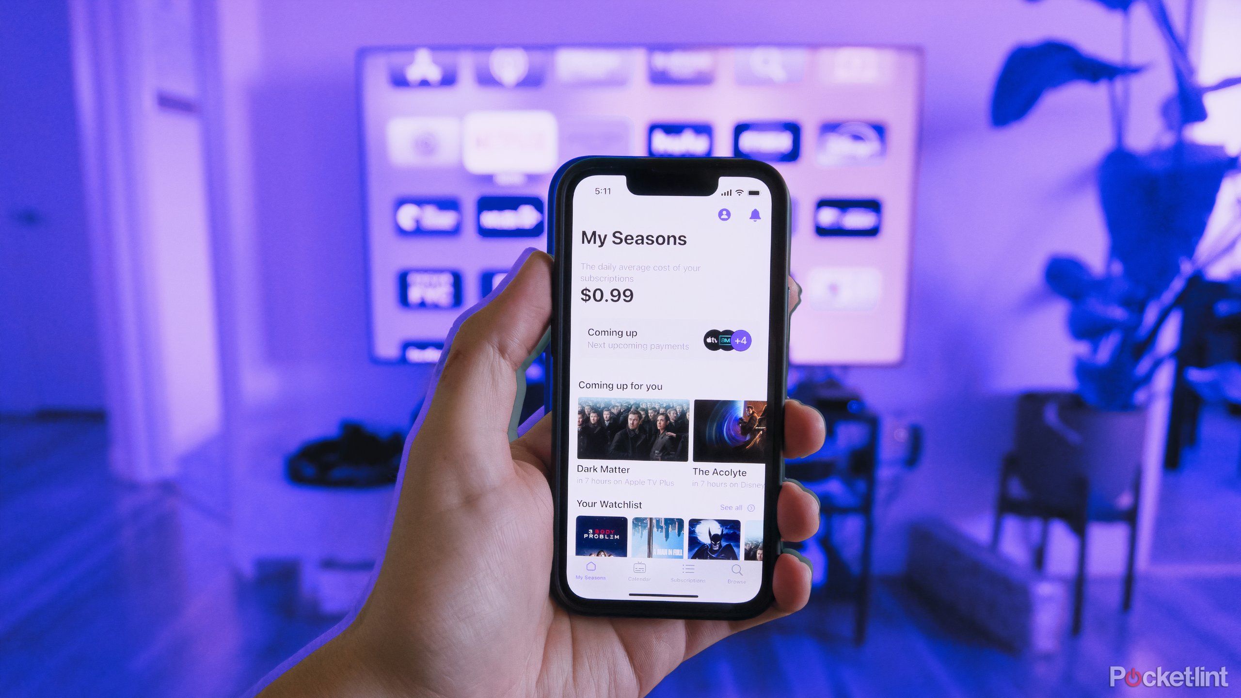 A hand holds an iPhone 13 mini displaying the Seasons app in front of a TV displaying streaming apps.