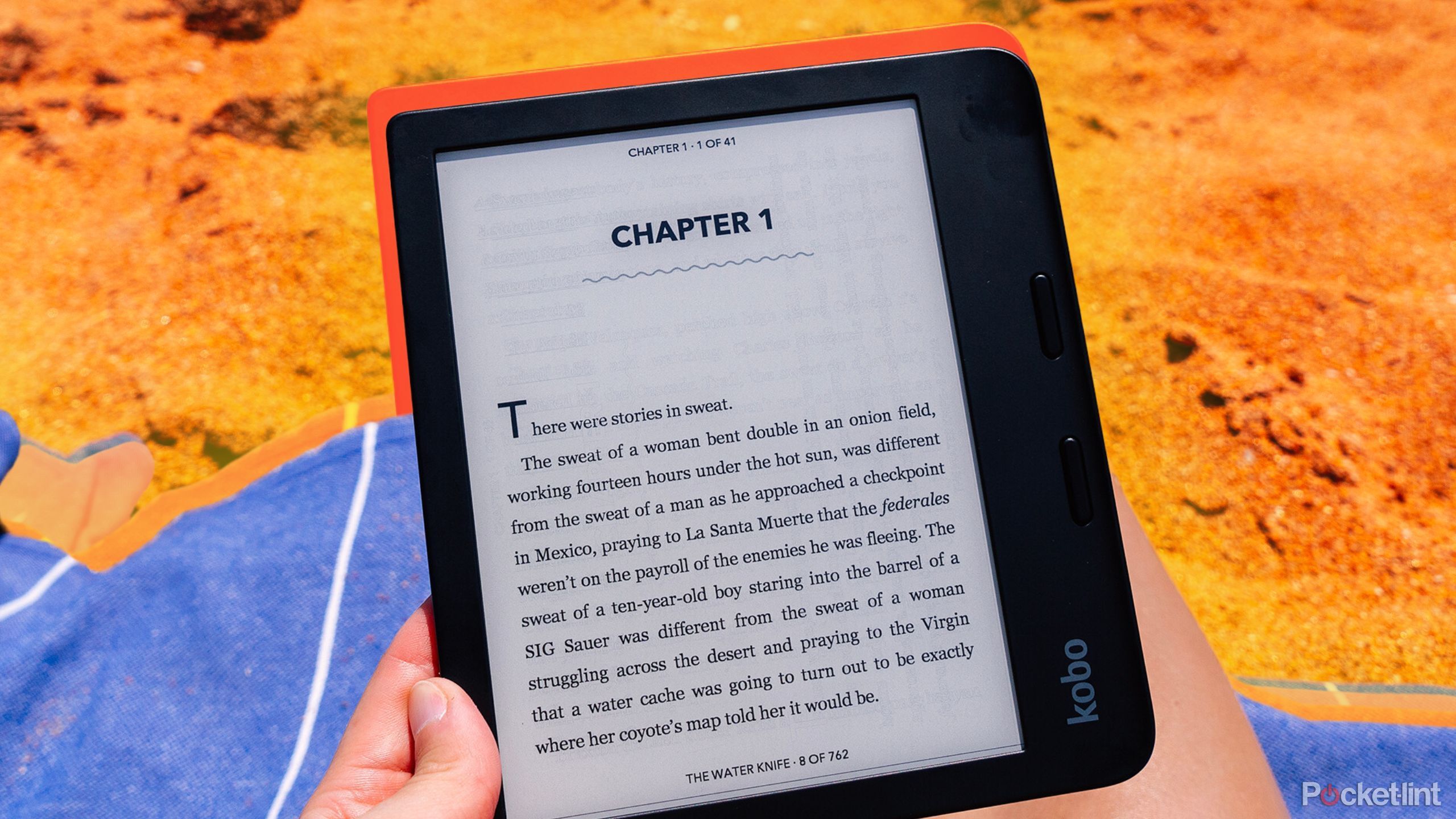 A hand holds the Kobo Libra 2 in front of a blue beach blanket on orange sand. 