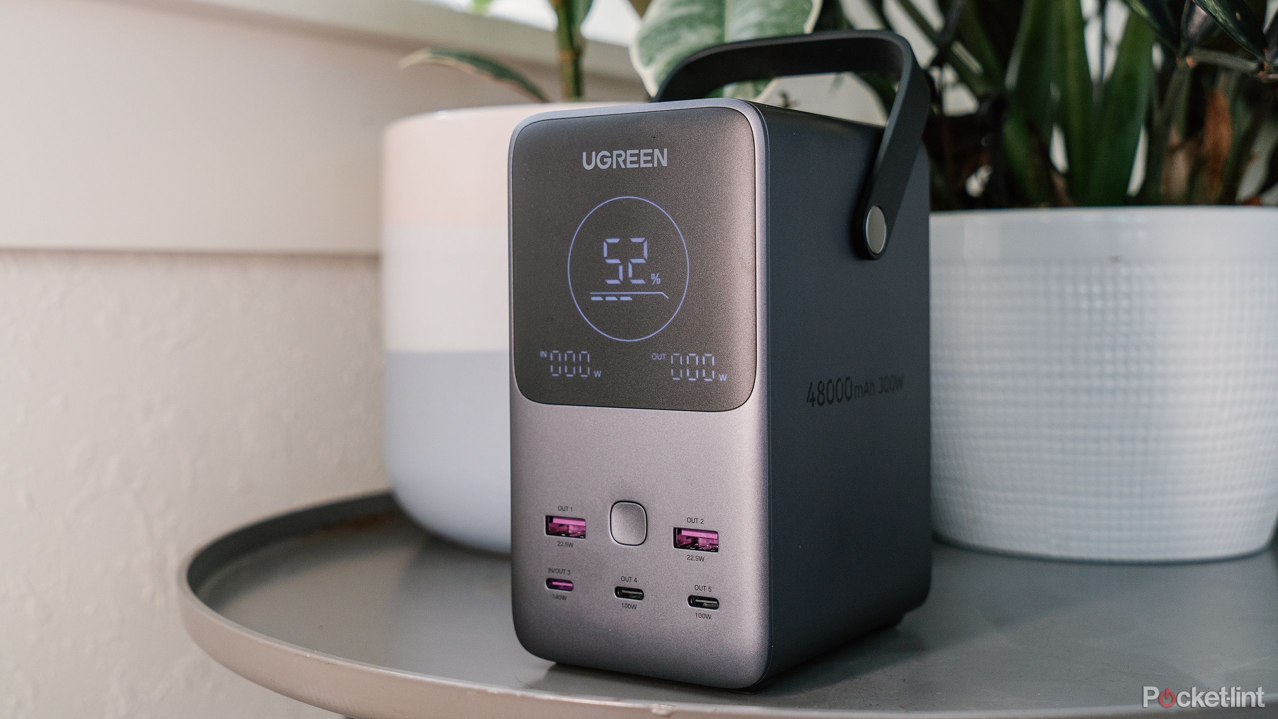 The Ugreen 300W 48000mAh Power Bank sits on a gray table in front of potted plants. 