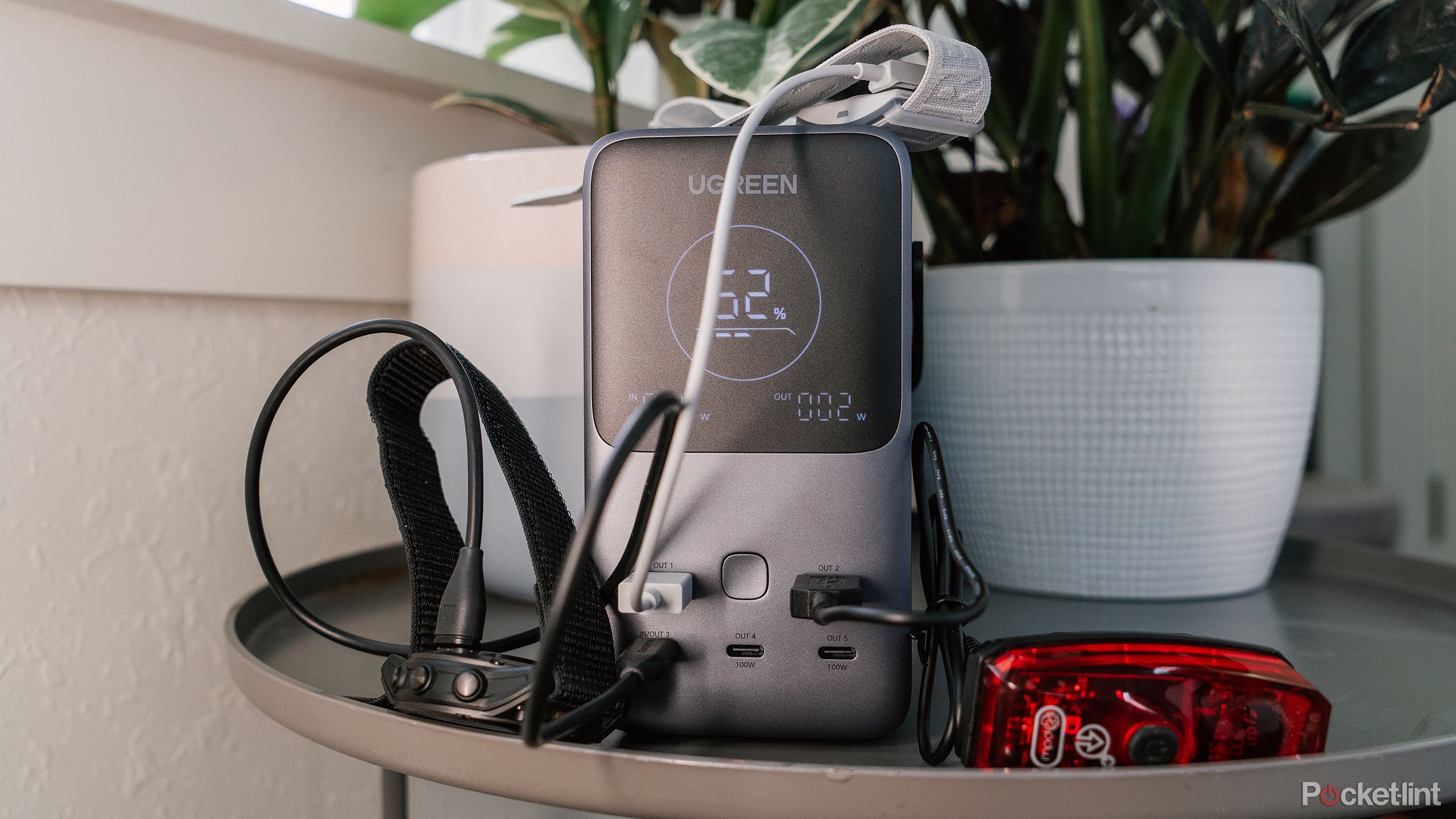 A pile of small devices and cords sit in front of the Ugreen 300W 48000mAh Power Bank on a gray table in front of potted plants. 