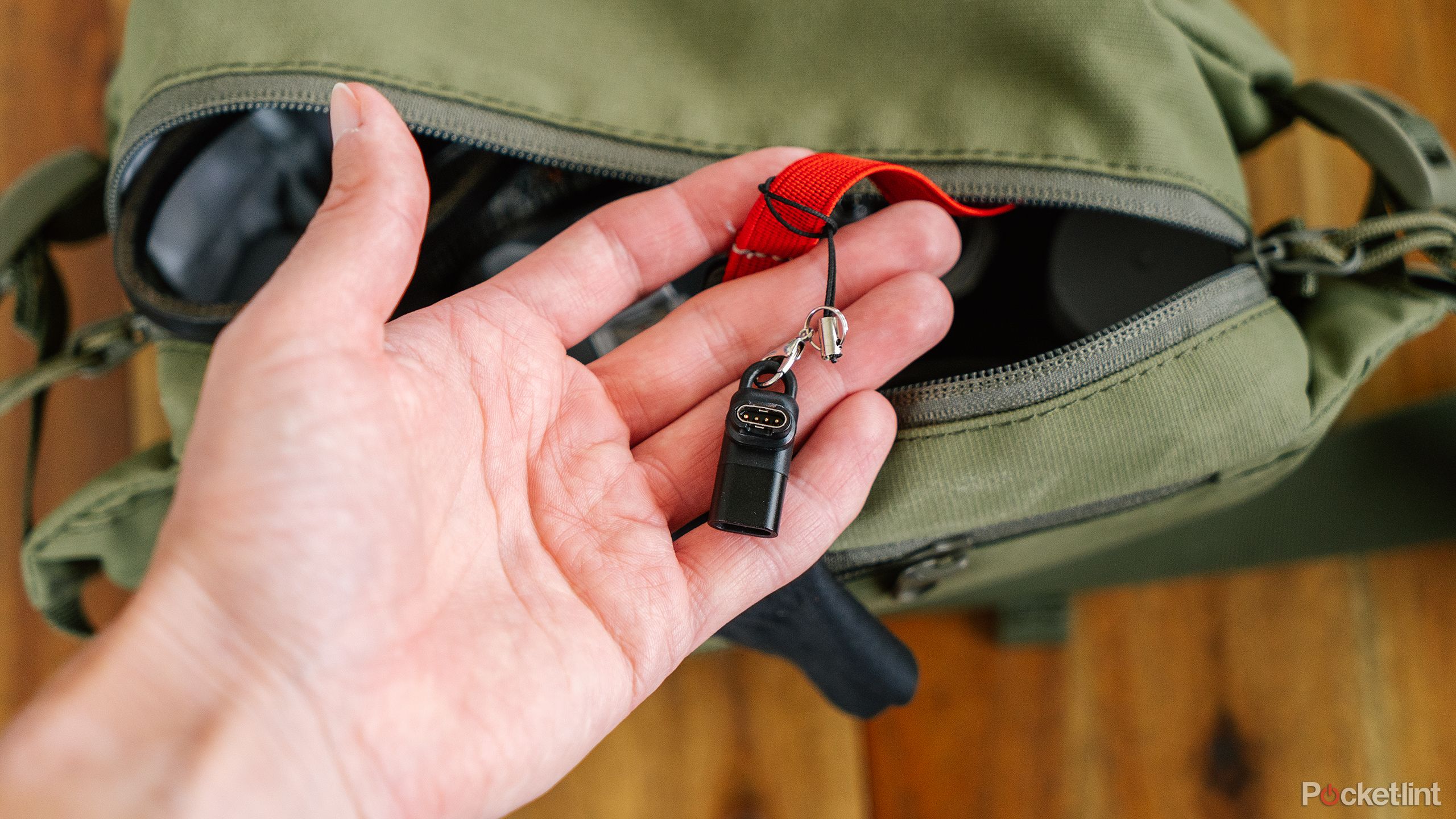 A hand holds up the Abanen Charging Adapter while it's attached to a red strap in a green bag. 