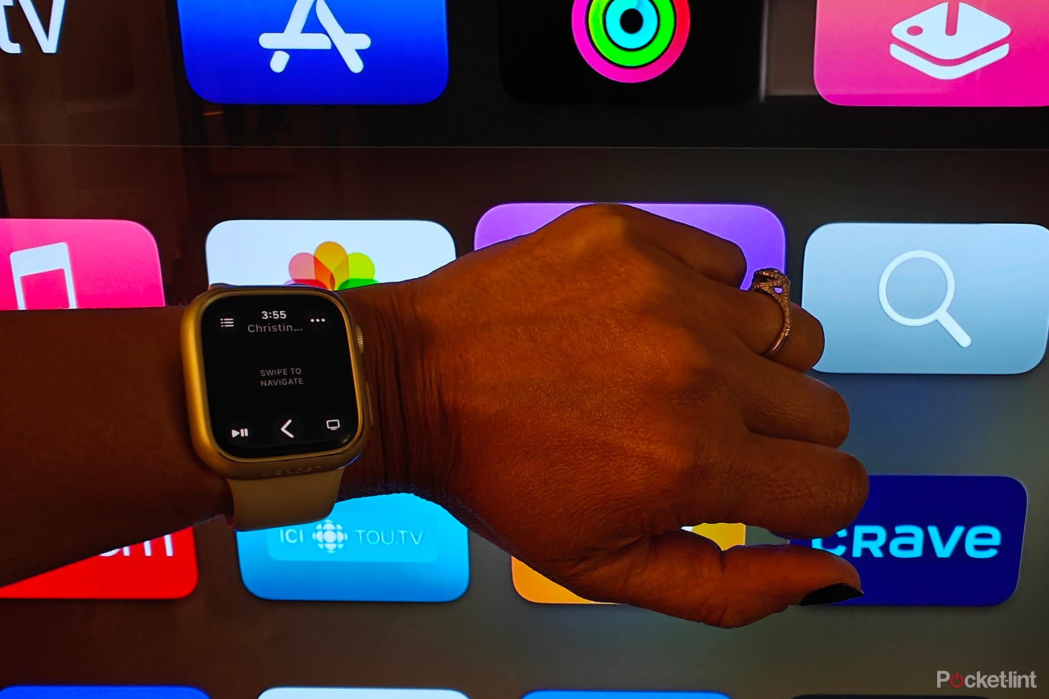 An Apple Watch being held up to a TV showing the Apple TV home screen, controls on the watch face.