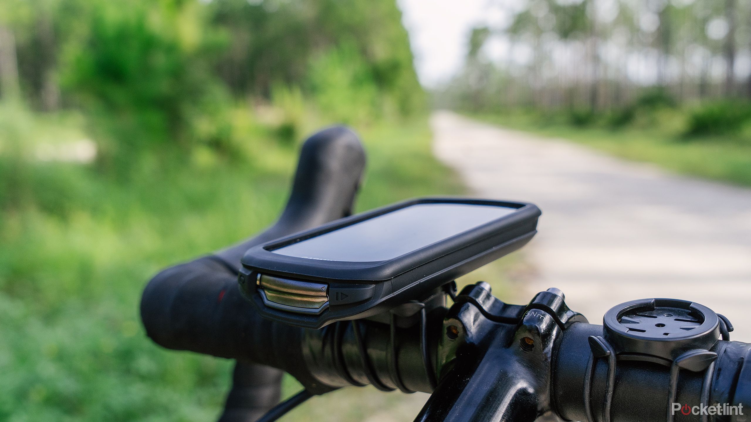 The Garmin Edge 1040 Solar is attached to bike handlebars in front of a blurred out gravel road and green forest. 