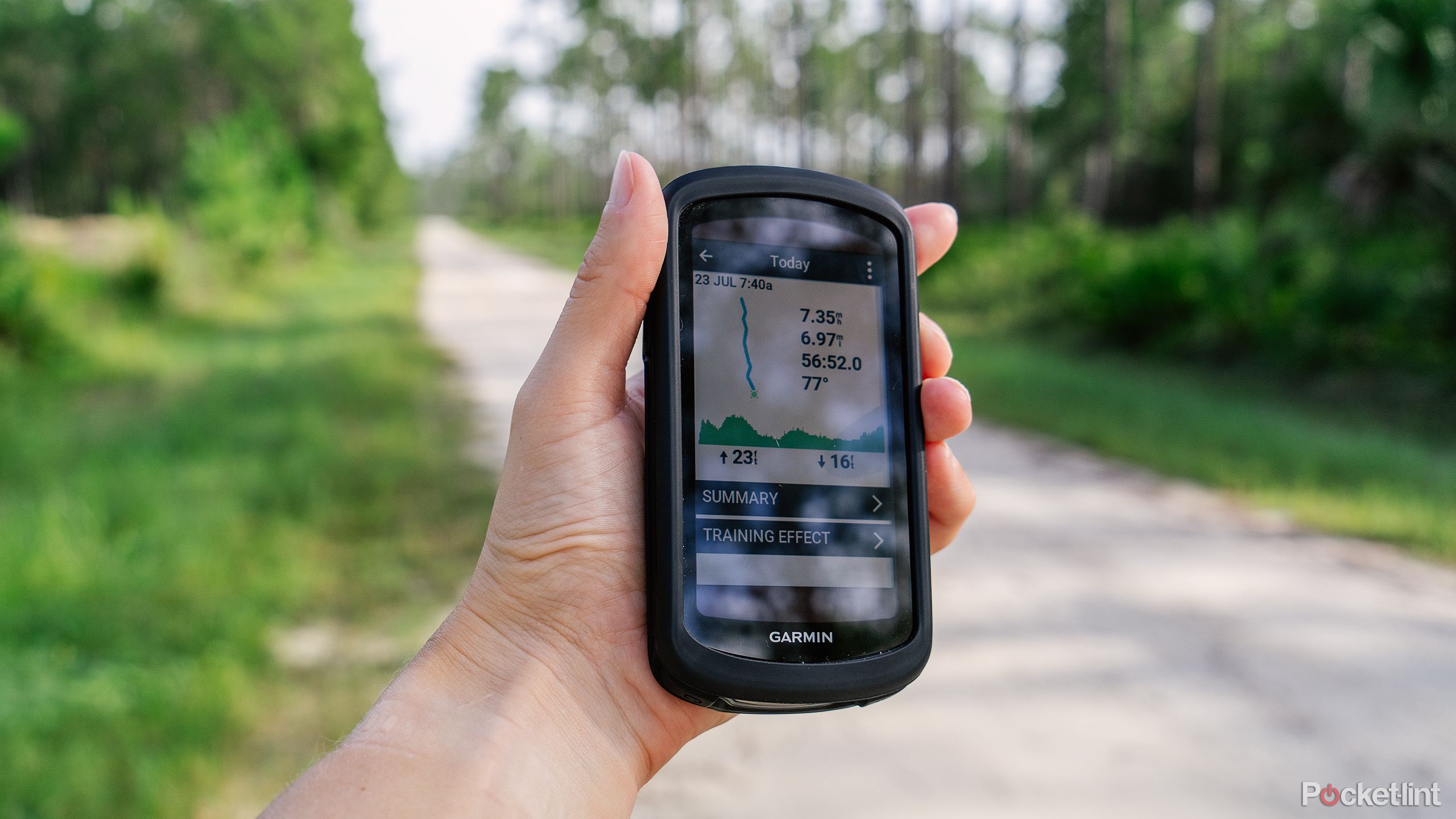 A hand holds the Garmin Edge 1040 Solar in front of a gravel road in a forest. 