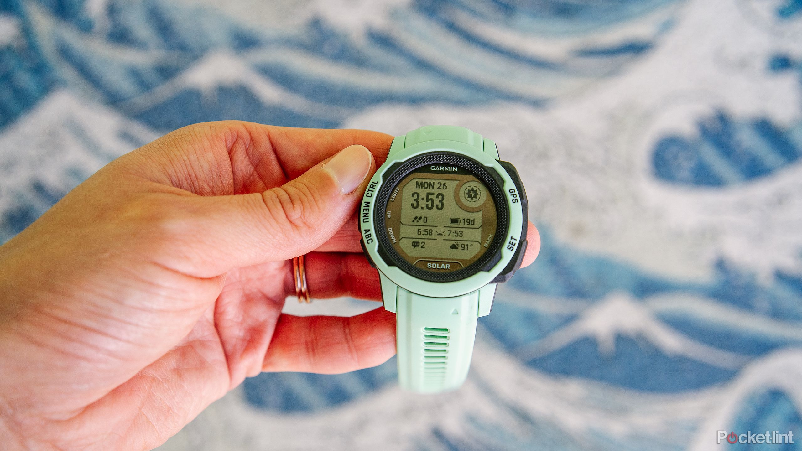 A hand holds the mint Garmin Instinct 2S above a blurred graphic of waves. 