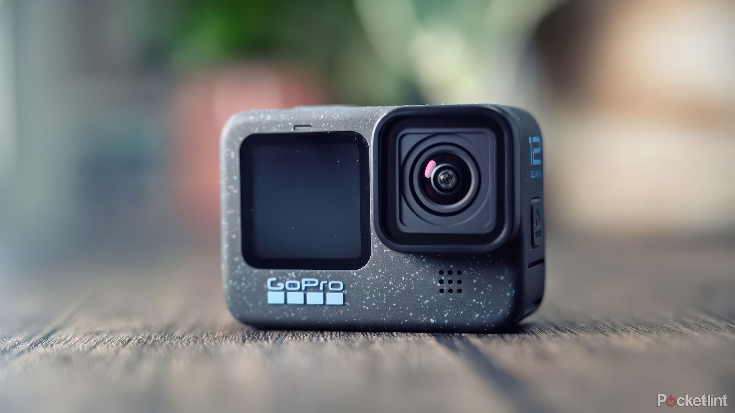 The GoPro Hero 12 Black sits on a wooden table in front of blurred plants. 