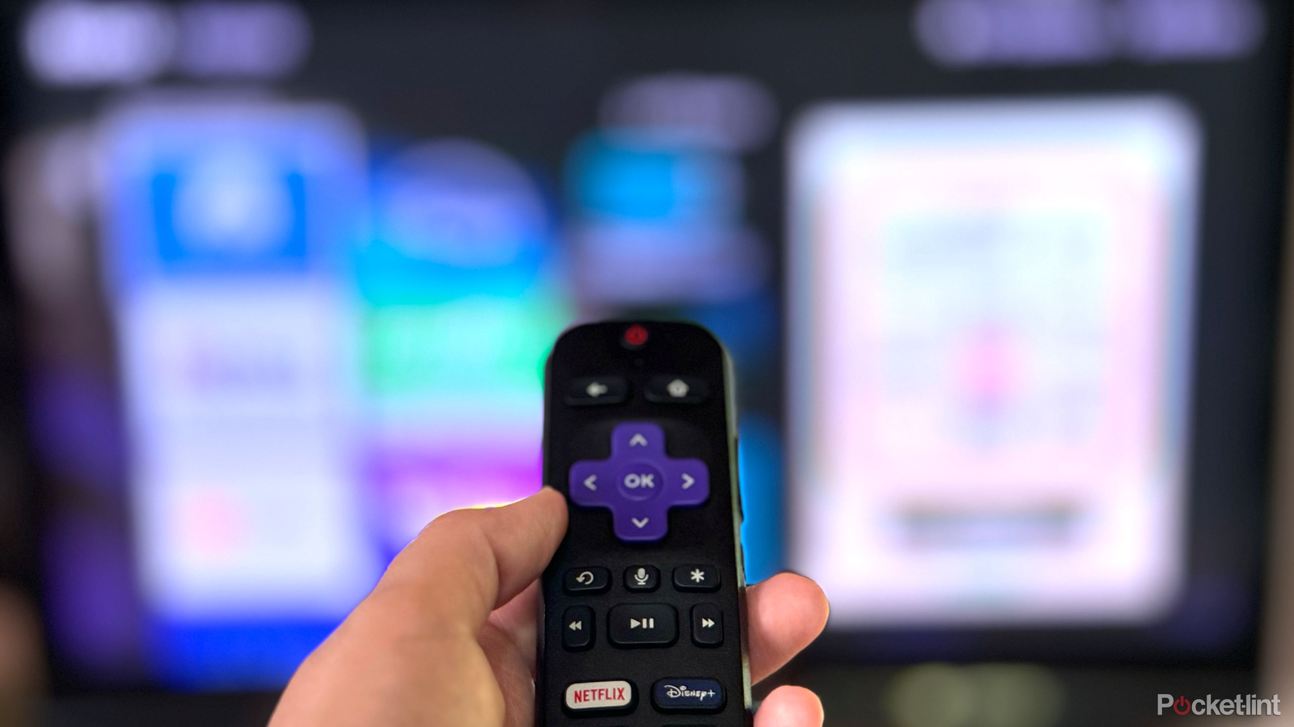 Uma foto de uma mão segurando um controle remoto Roku na frente de uma televisão