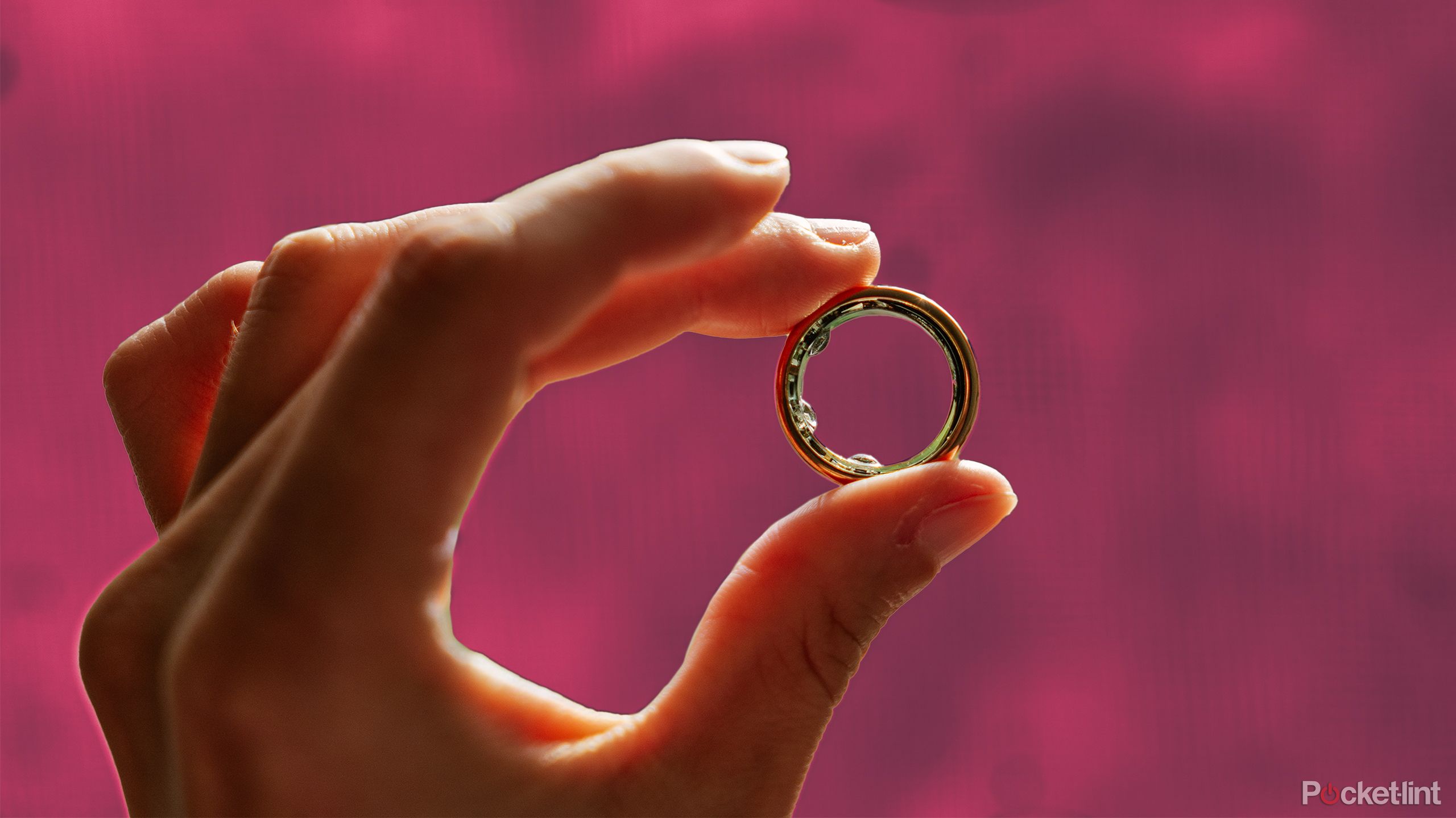 A hand holds the Oura Ring between its fingers in front of a dark pink background. 