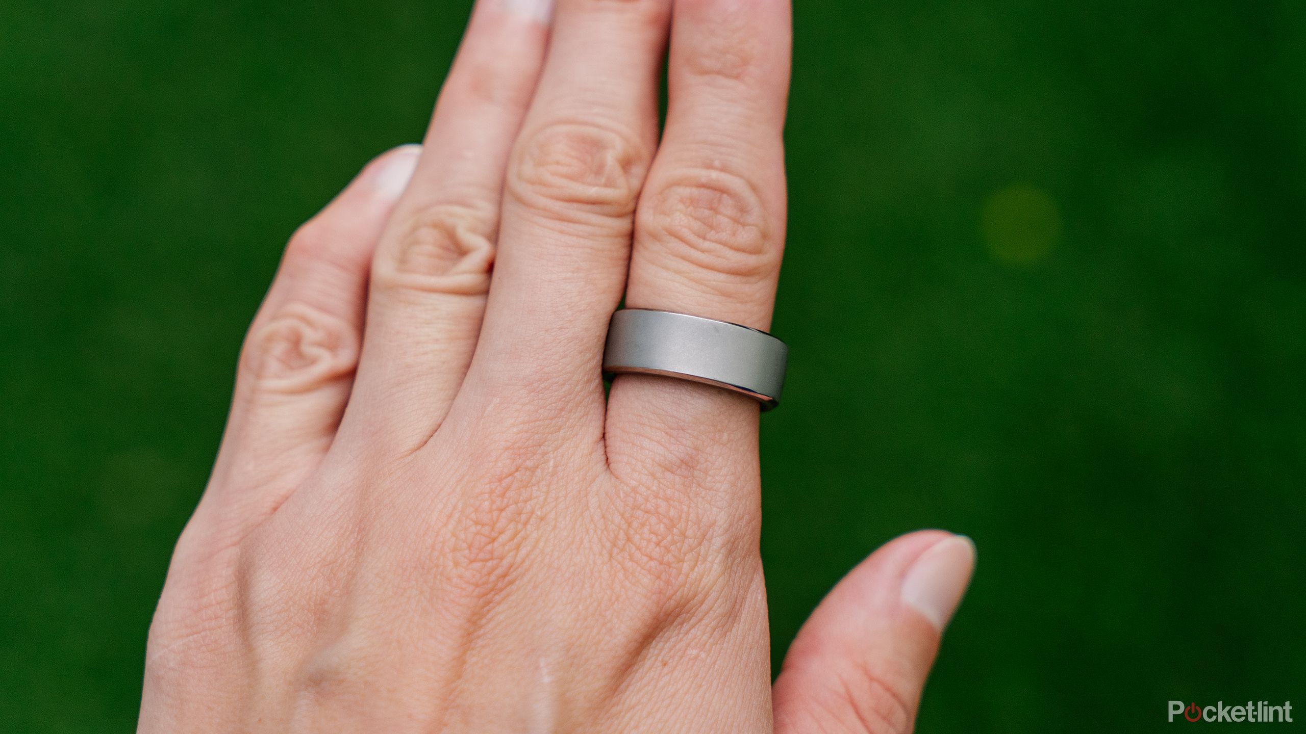 A hand is held out over a blurred green background with the RingConn Smart Ring on the index finger. 