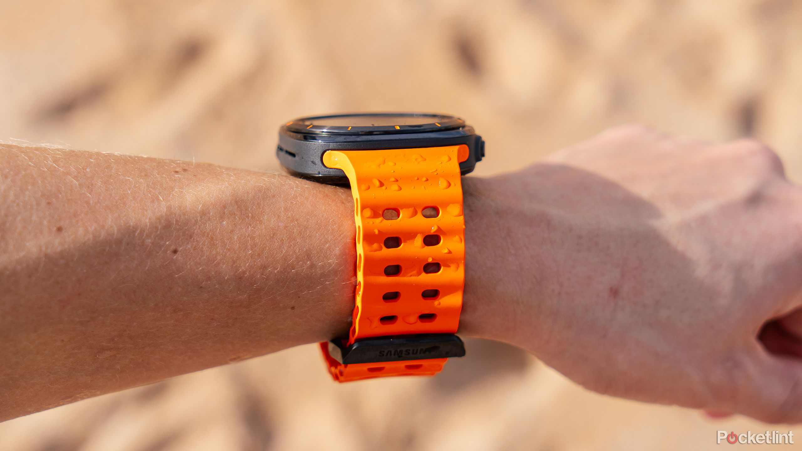 The Samsung Galaxy Watch Ultra is on a wrist held above blurred out sand. 