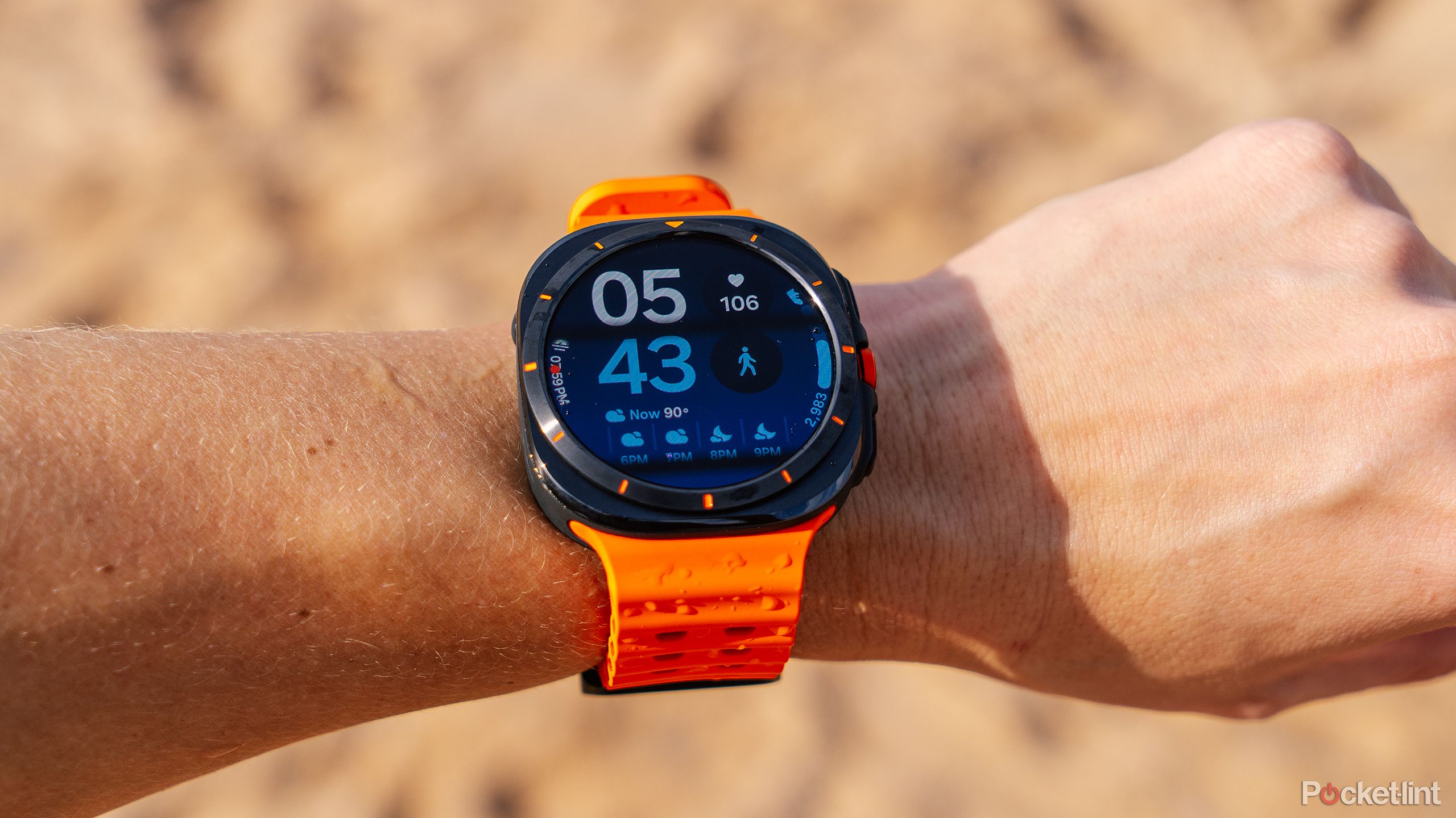 The Samsung Galaxy Watch Ultra is attached to a wrist held in front of a blurred beach. 