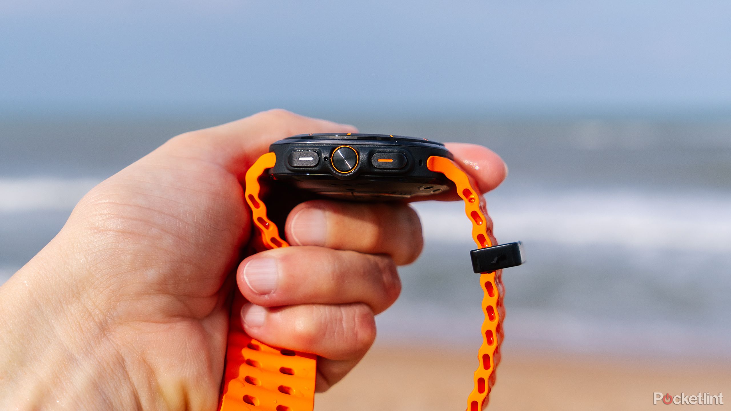 A hand holds the Samsung Galaxy Watch Ultra in front of a blurred out beach. 