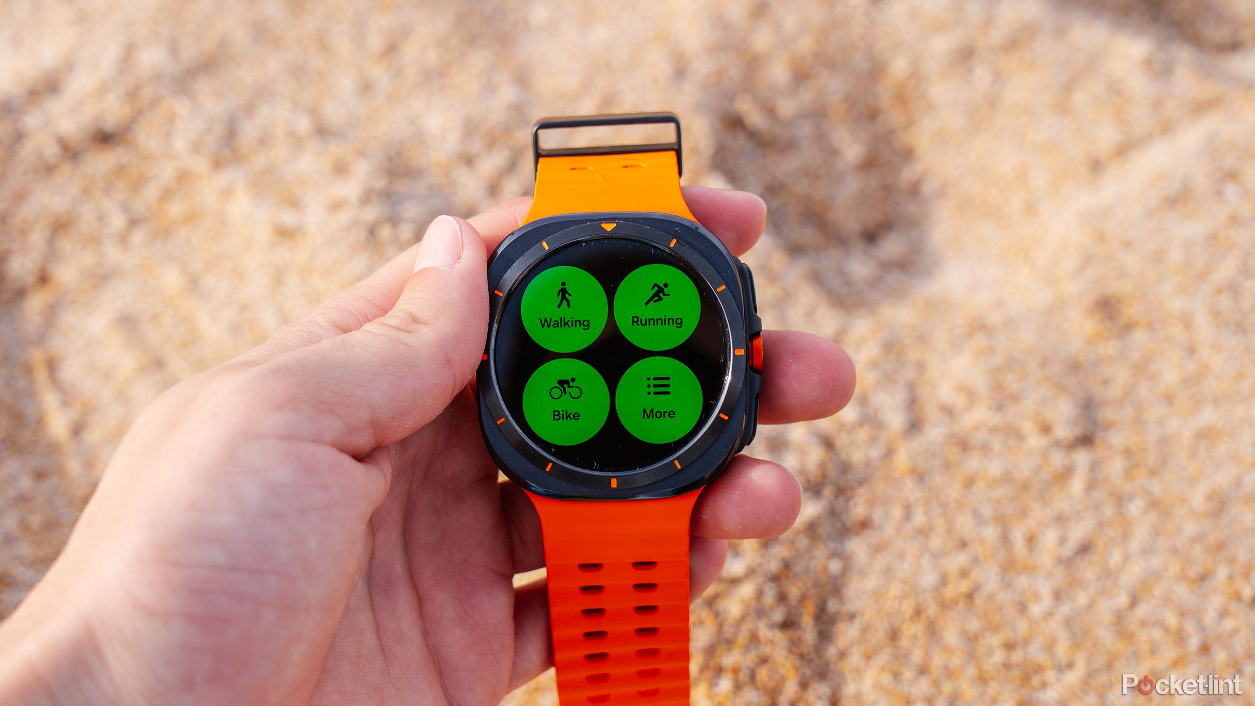 A hand holds the Samsung Galaxy Watch Ultra over sand. 