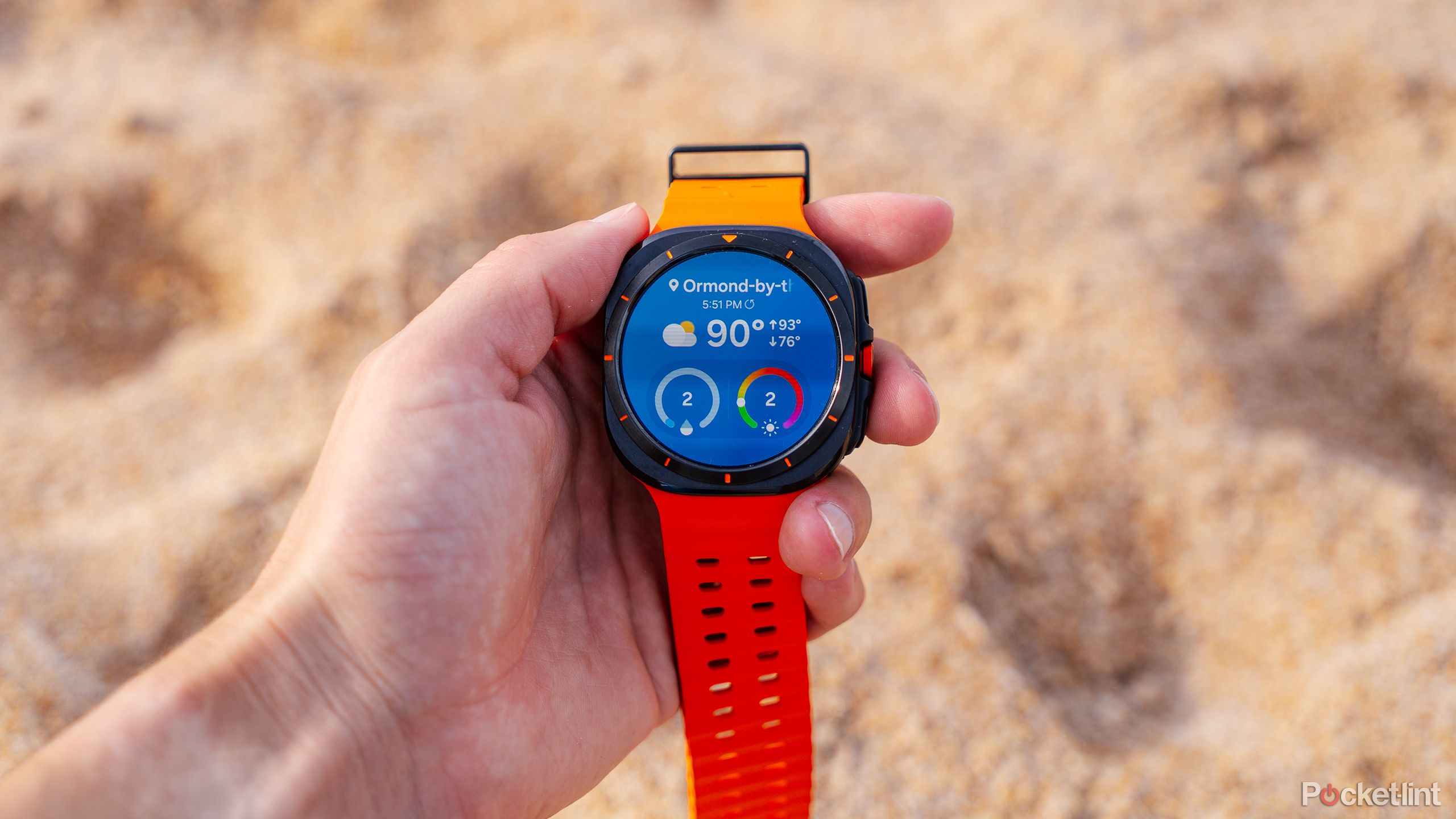 A hand holds the Samsung Galaxy Watch Ultra over sand. 