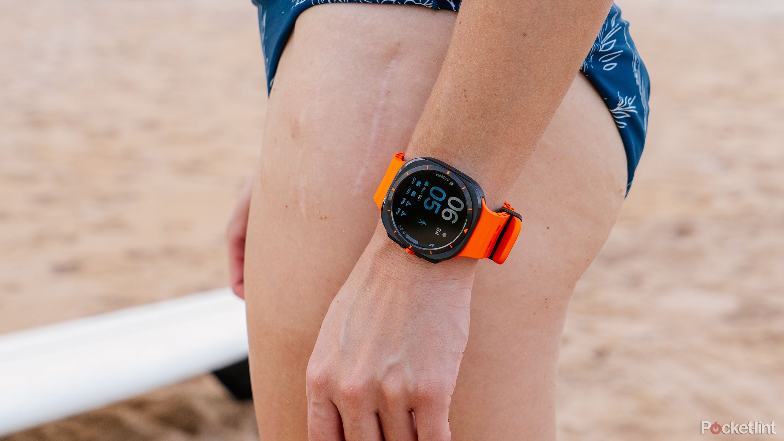 The Samsung Galaxy Watch Ultra is on a wrist that rests against a person's leg while standing on the beach.