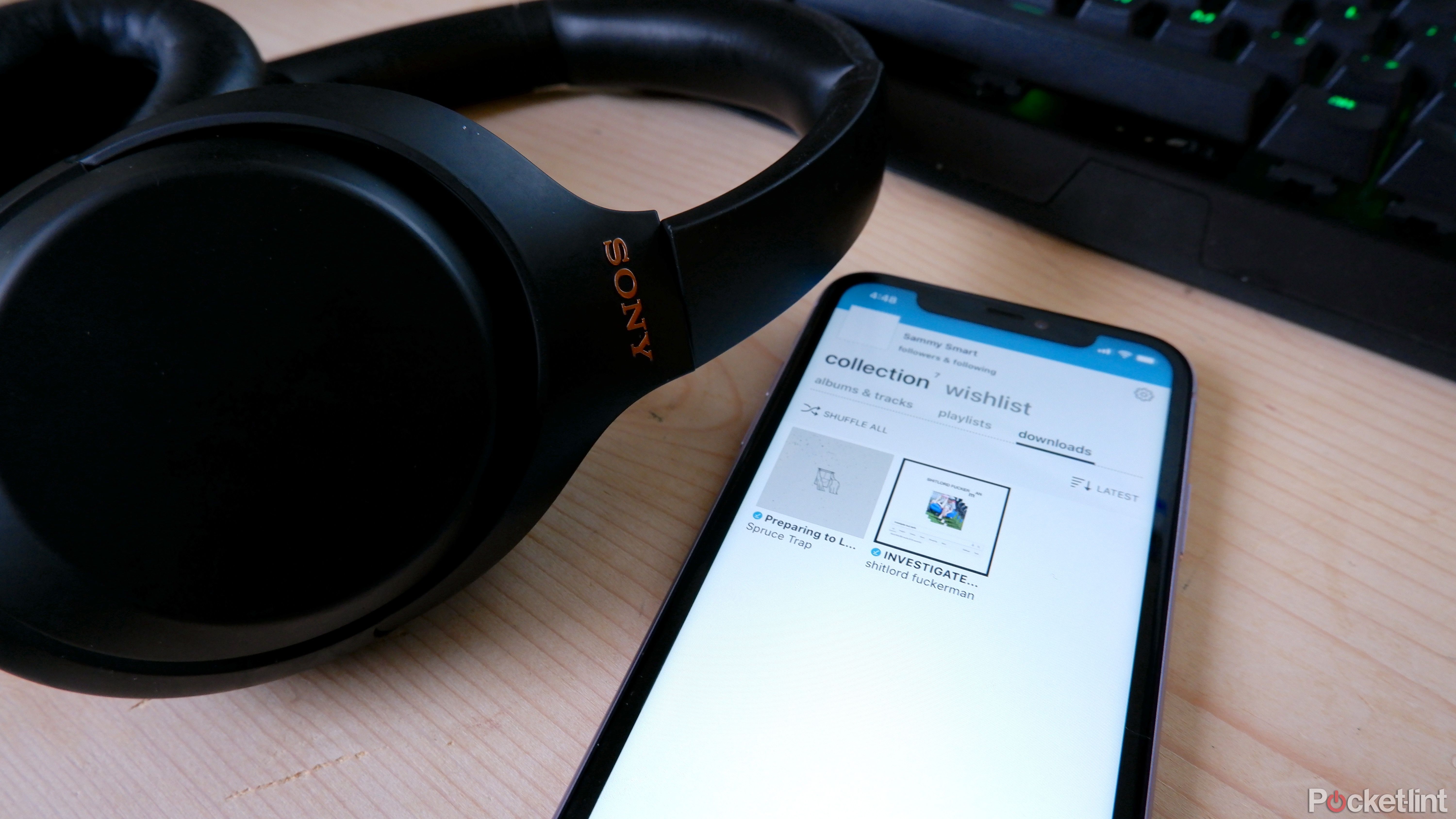 An iPhone on a wooden table with the Bandcamp open showing downloaded music, next to a pair of Sony WH-1000XM4 headphones