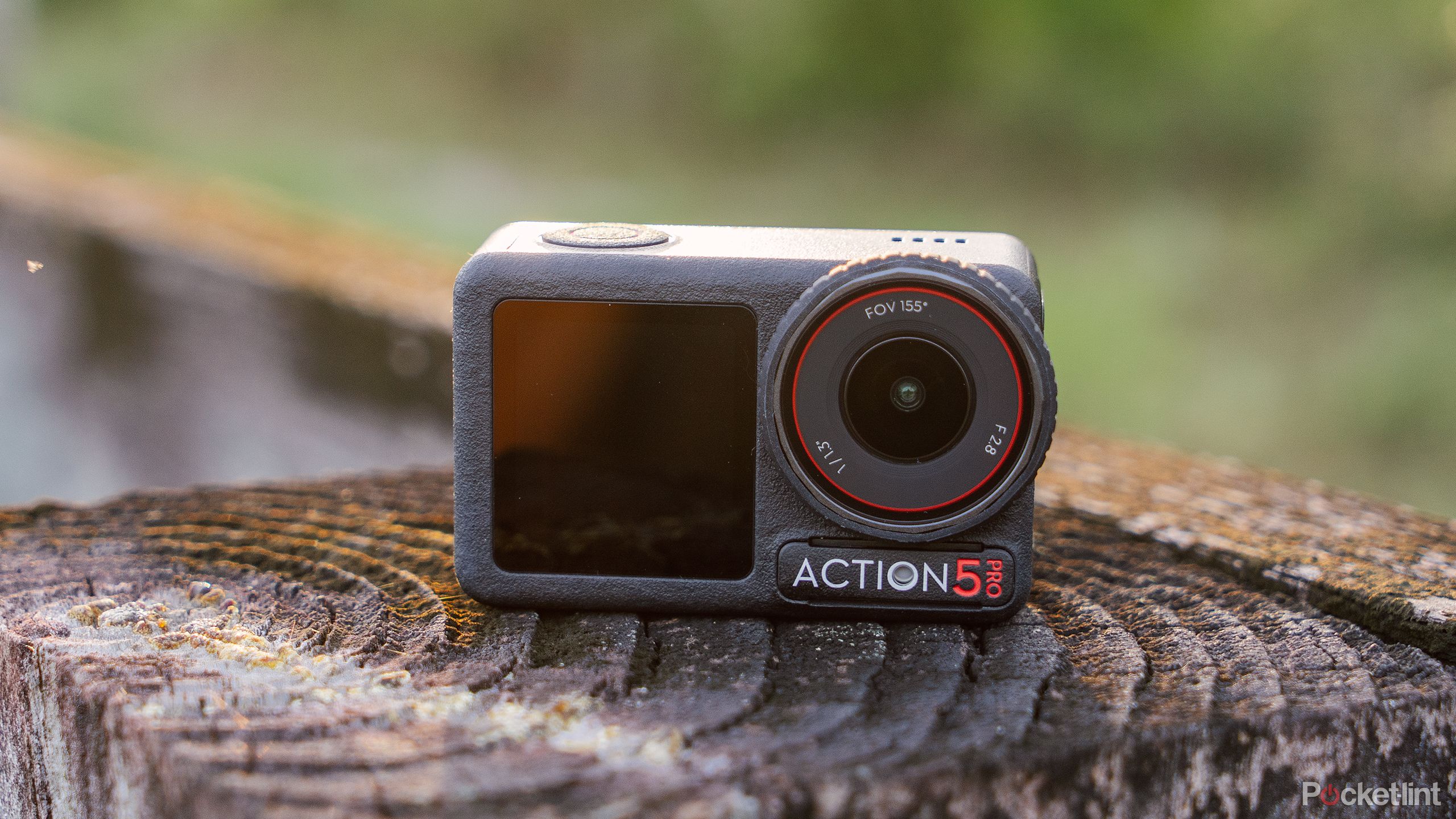 The DJI Osmo Action 5 Pro sits on a weathered fence post in front of a blurred green forest. 