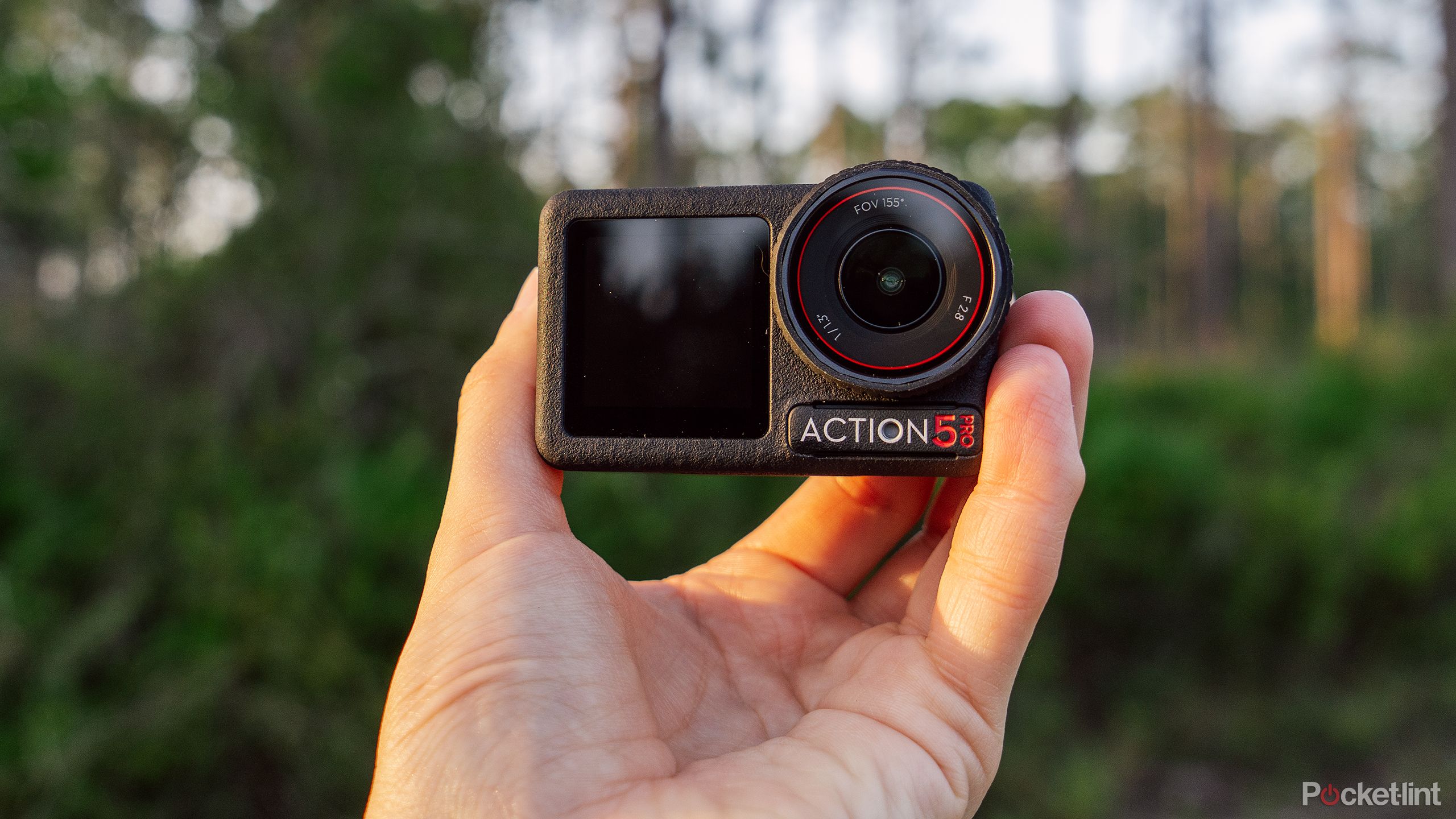 A hand holds the DJI Osmo Action 5 Pro in front of a blurred forest. 